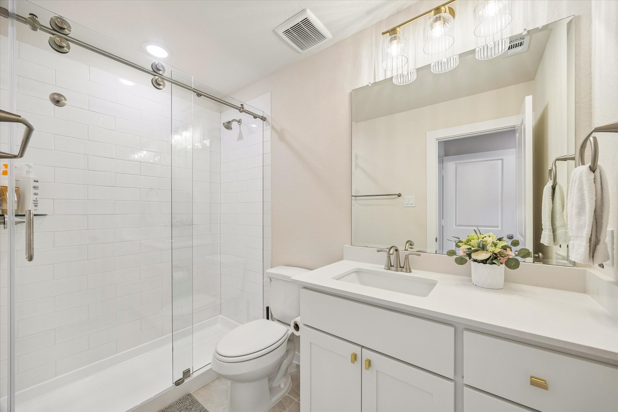 SECONDARY BATH FEATURES A GLASS-ENCLOSED SHOWER, WHITE TILE SURROUND, QUARTZ VANITY, AND MODERN LIGHTING.
