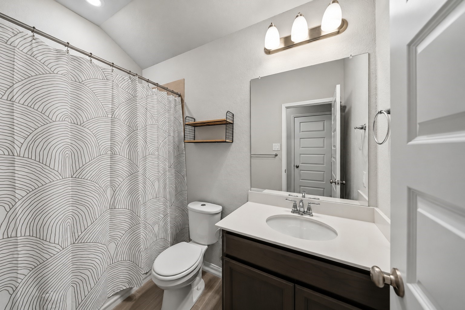 Virtually Enhanced: This well-appointed bathroom features a clean, functional layout with soft neutral walls and durable wood-look flooring. A spacious single-sink vanity offers a white countertop, an undermount basin, and dark cabinetry for convenient storage. A large wall-mounted mirror, three-light vanity fixture, and recessed ceiling lighting provide excellent illumination.
