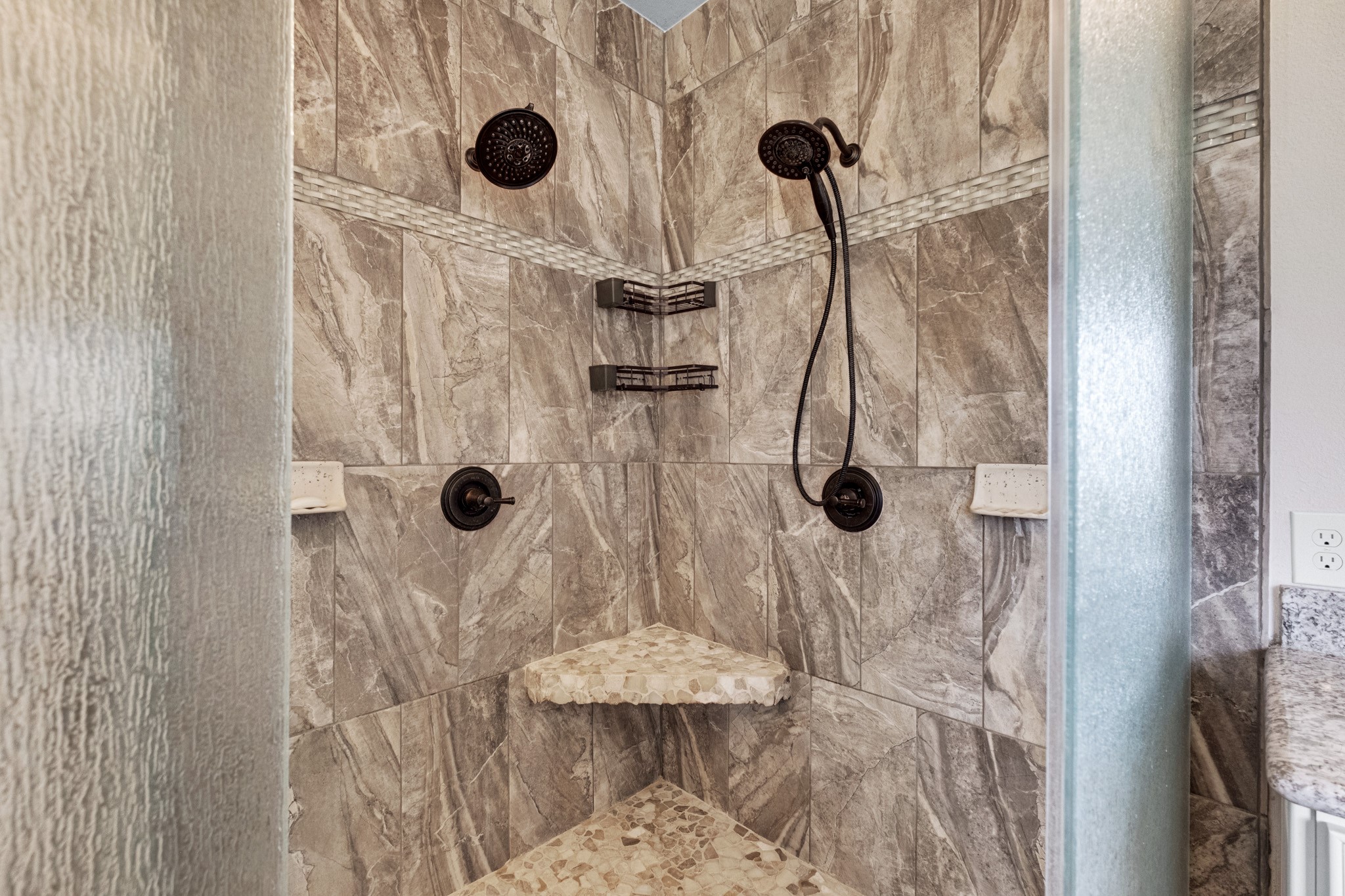 The walk-in shower is finished floor to ceiling in stone-look tile with dual shower heads, a handheld on slide bar, corner seat, and pebble mosaic floor.