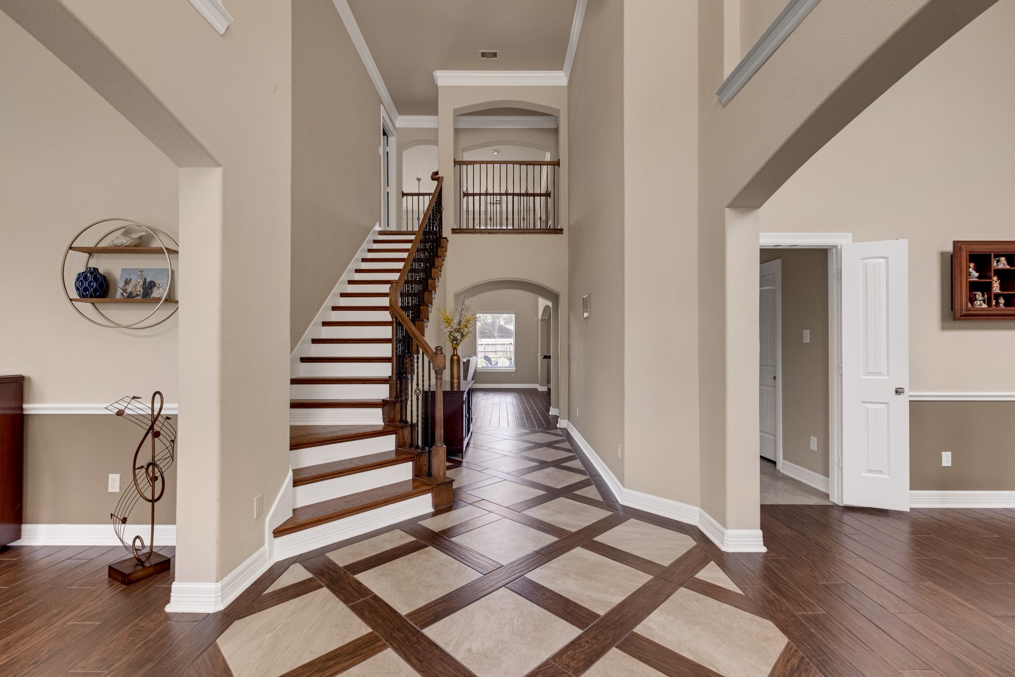 Step inside to a two-story foyer laid in a harlequin inlay - travertine set on point, ribboned in mahogany wood tile - with a hardwood staircase and wrought-iron balusters climbing to the railed catwalk above, crown molding and chair rail carrying the formality down the hall.