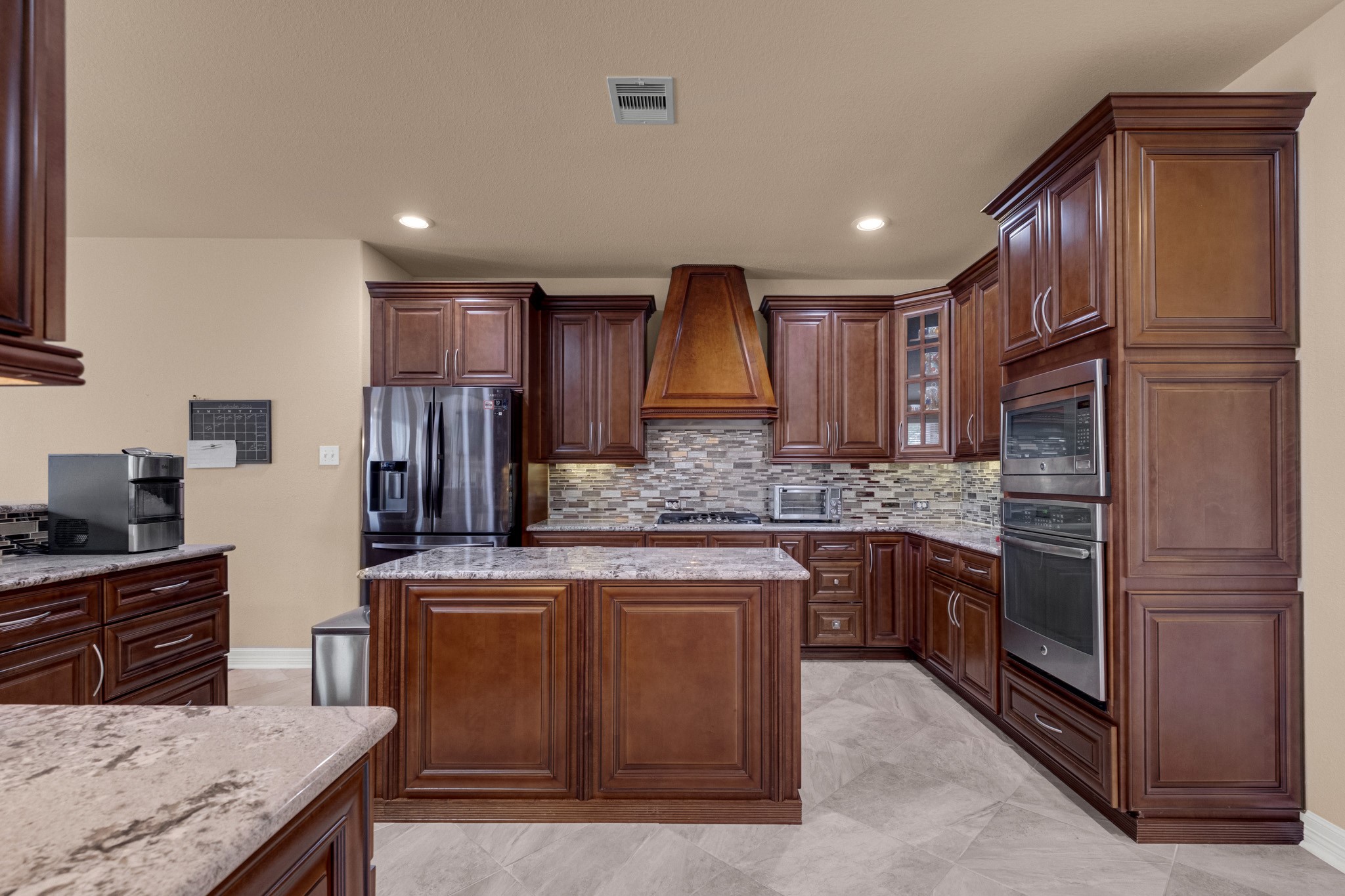 The kitchen is dressed in chocolate-glazed cherry cabinetry with a custom tapered wood chimney hood, full-movement granite counters, a linear glass and travertine mosaic backsplash, and a stainless wall oven with microwave above, set into a full-height cabinet tower.