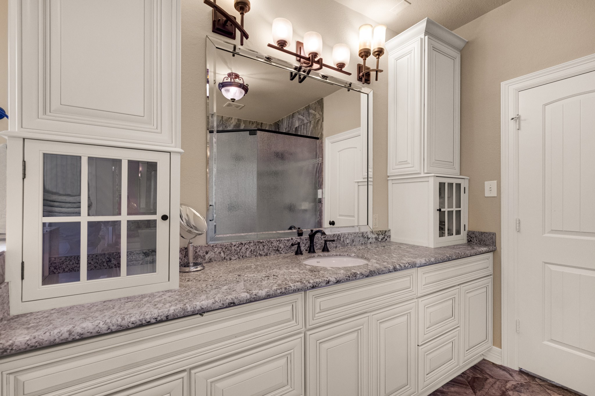 The vanity wall stretches long in granite with framed mirror, sconce lighting, and glass-front cabinetry for display and storage together.