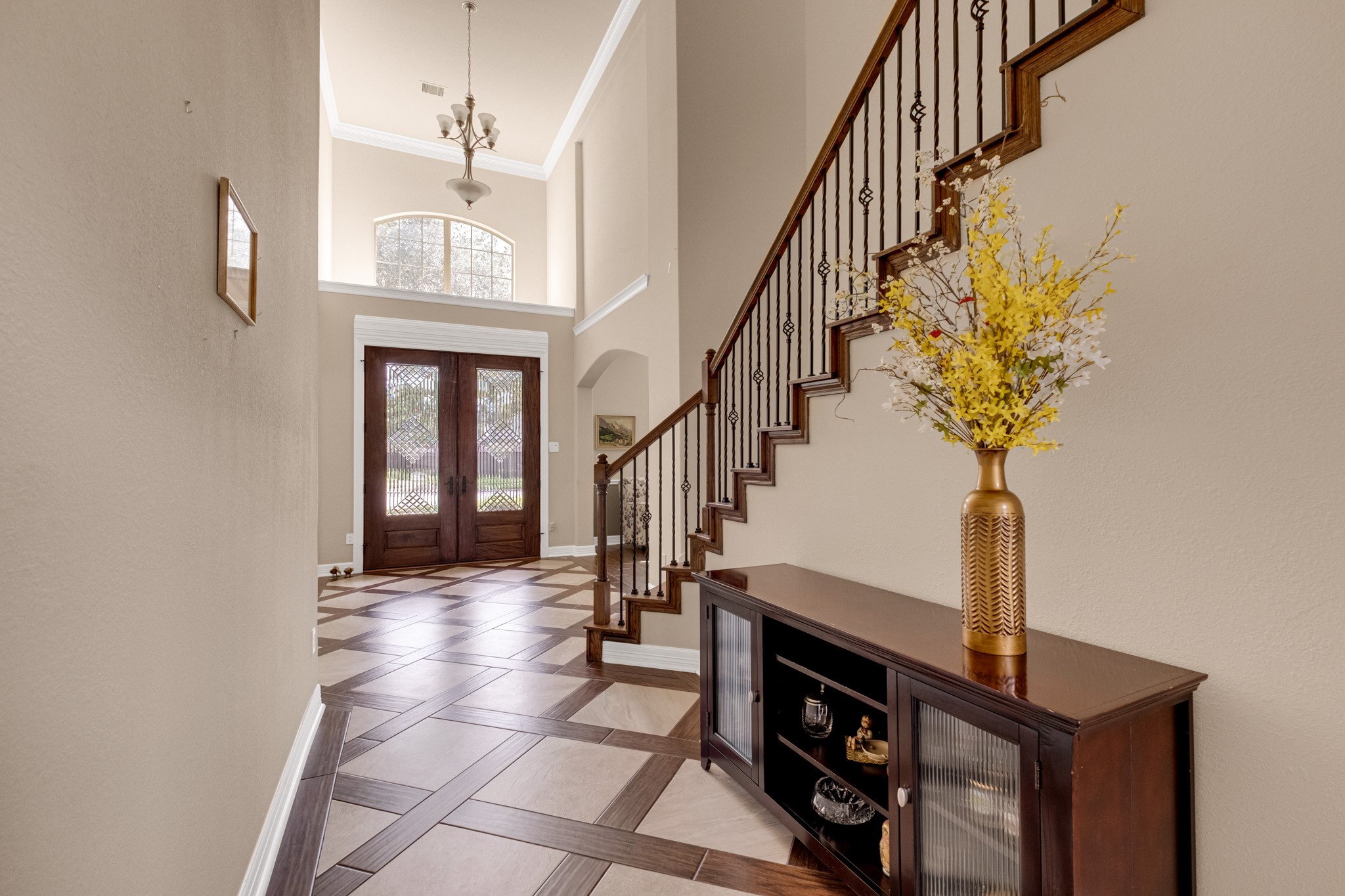 Looking back toward the entry, an arched transom window floats above the doors and a chandelier hangs the full two stories, with the carved newel post and iron balusters anchoring the run.
