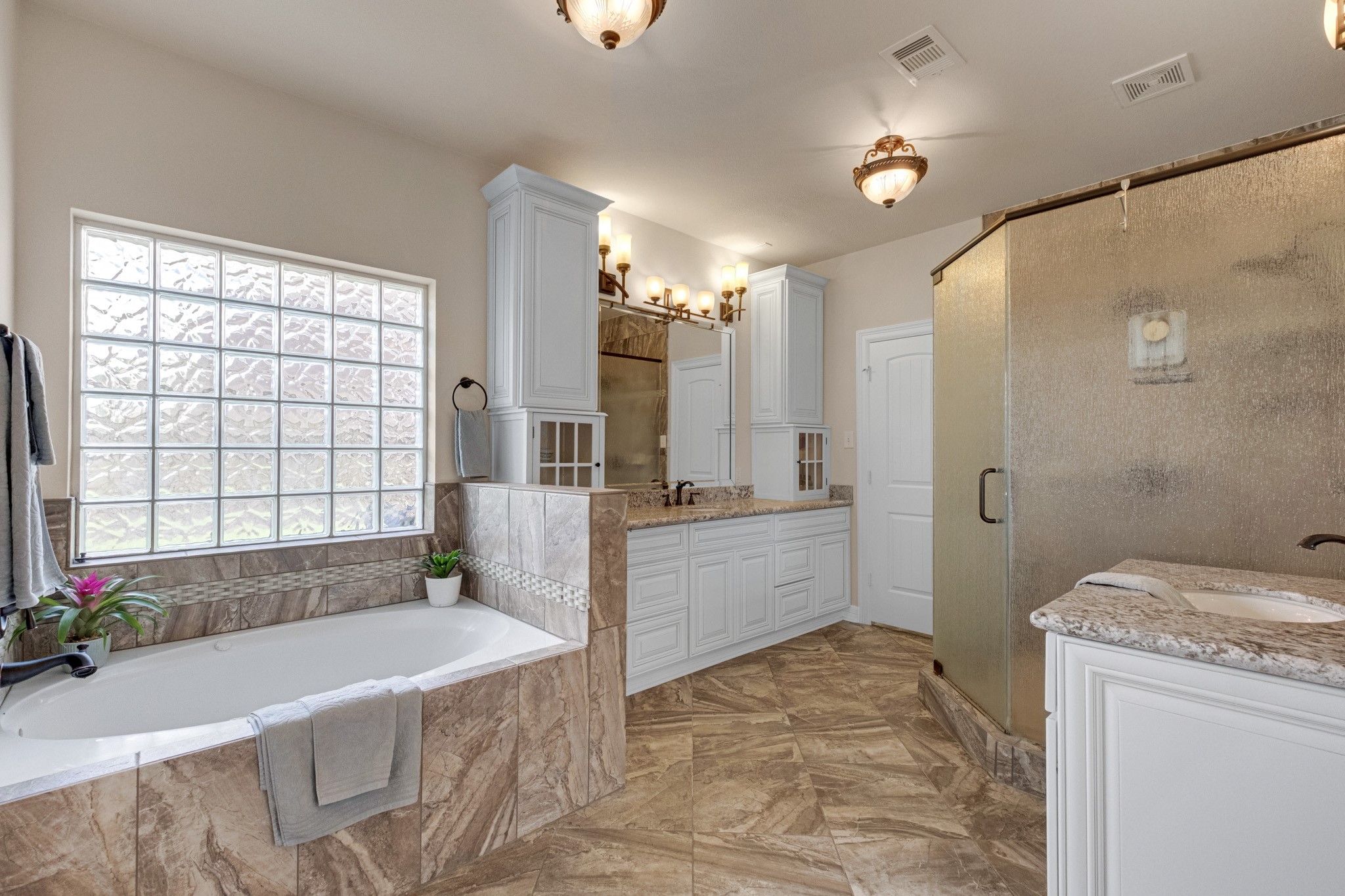 The primary bath is a full retreat: a tiled garden tub beneath a glass block window, dual granite vanities with framed mirrors, and a separate glass-enclosed shower.