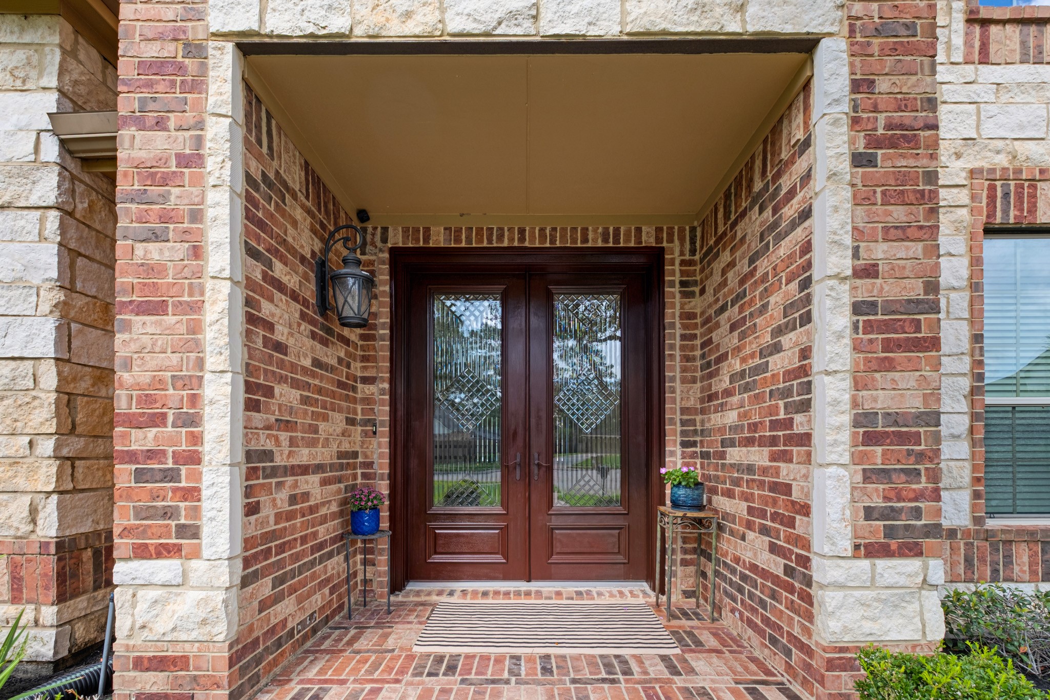 The covered entry frames a mahogany-toned double door set with wrought-iron diamond-lattice glass, standing between cast stone quoins and running-bond brick, with a herringbone brick porch underfoot.
