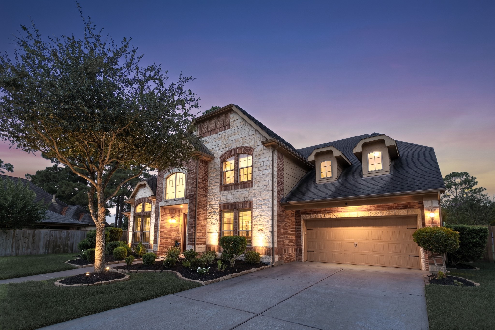 Arrive at dusk on a quiet Bayou Park 3 cul-de-sac and the elevation does the talking - chopped limestone masonry set against contrast red brick, gabled dormers catching the last of the light, and landscape uplighting washing the stonework while a mature oak shades the manicured lawn edged in stacked stone beds.