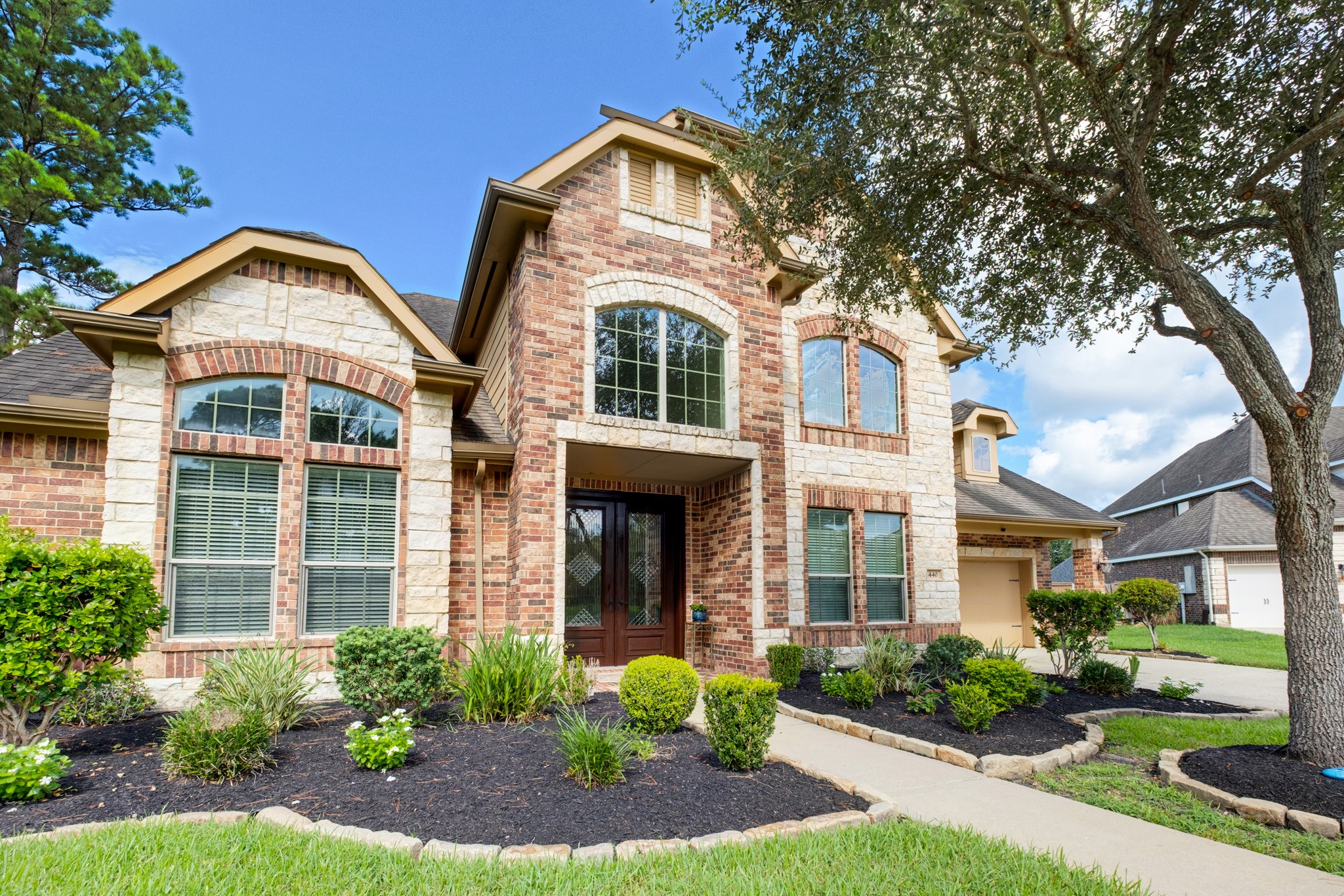 By daylight the craftsmanship reads even clearer: arched windows crowned with soldier-course brick and cast stone surrounds, a stained double entry door set with wrought-iron diamond-lattice glass, and a curved walkway winding through layered beds to the front step.