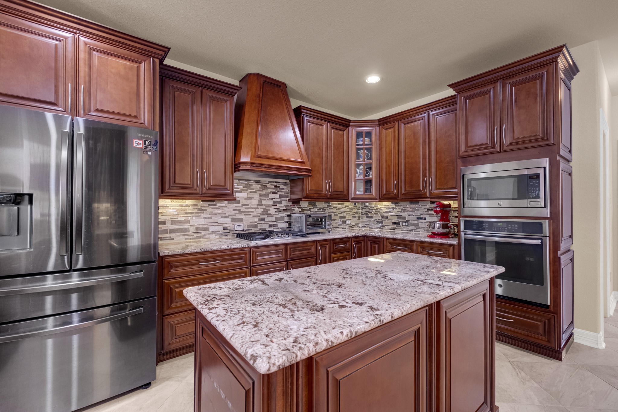The custom tapered wood chimney hood crowns a gas cooktop set in full-movement granite, with a stainless microwave and wall oven stacked in the cabinet tower and a French-door refrigerator alongside.