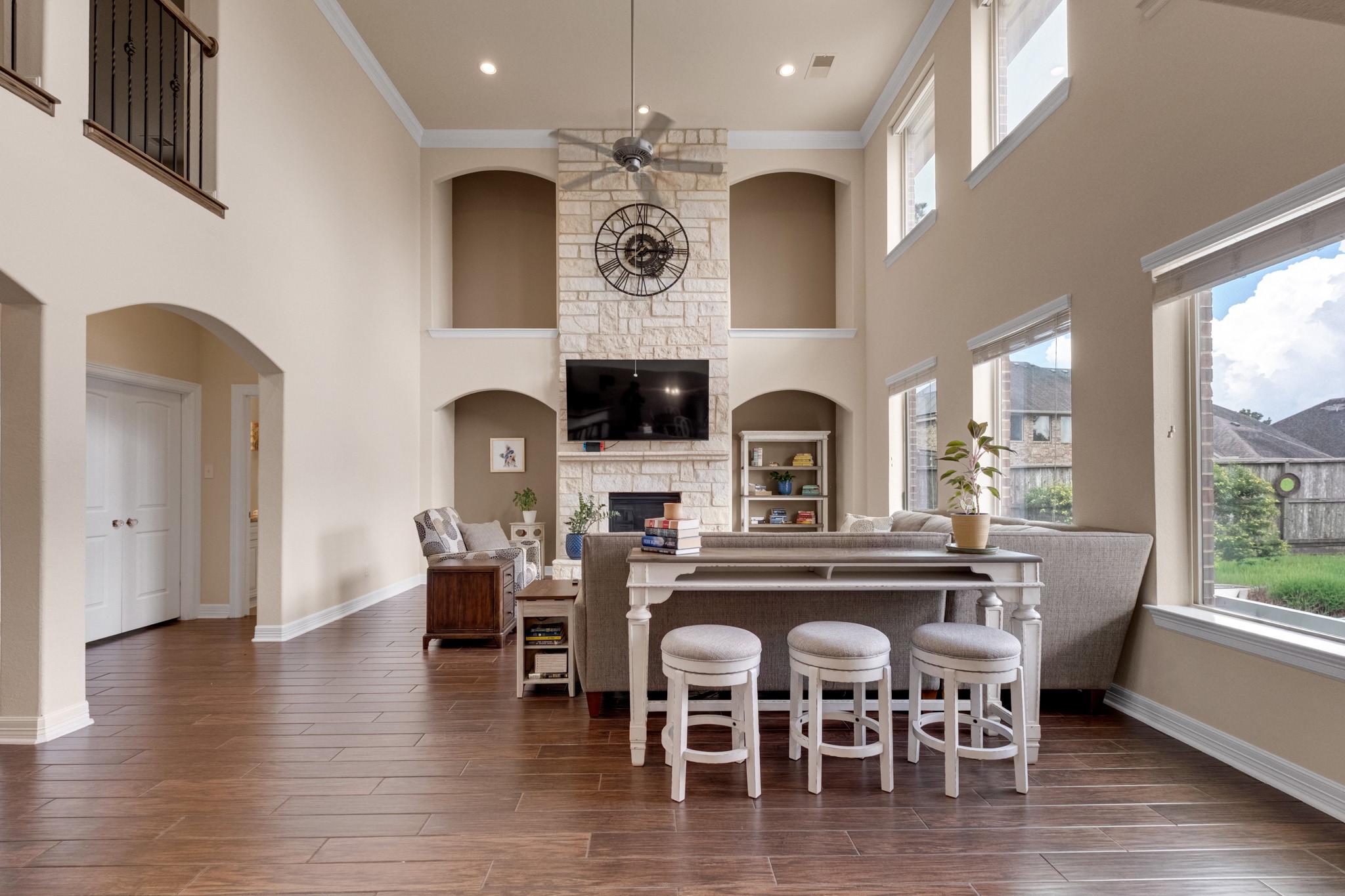 The family room opens to soaring two-story ceilings, anchored by a floor-to-ceiling dry-stack Austin stone fireplace flanked by arched display niches, with a wall of windows pouring light across mahogany wood tile.