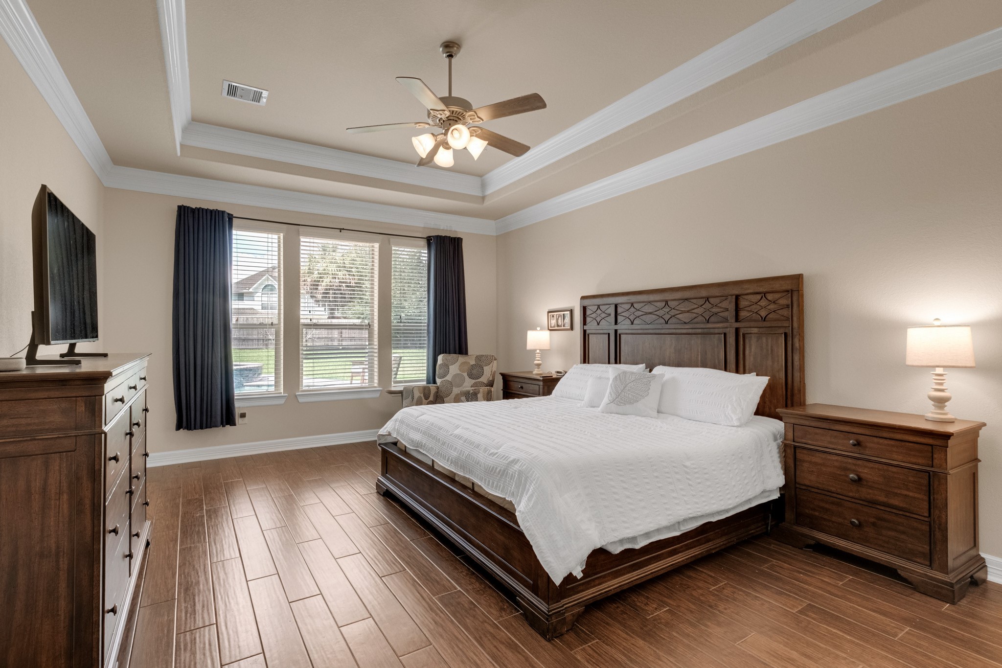 The primary suite retreats behind a stepped tray ceiling trimmed in crown molding, with mahogany wood tile underfoot and a wide window bank framing the shaded backyard.