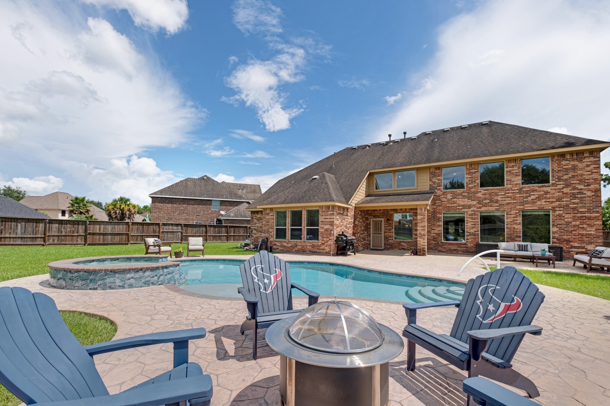 The pool comes into its own from the deck - freeform water with a raised stone spa spilling in, a fire pit ring in stamped concrete, and the two-story brick elevation as the backdrop.