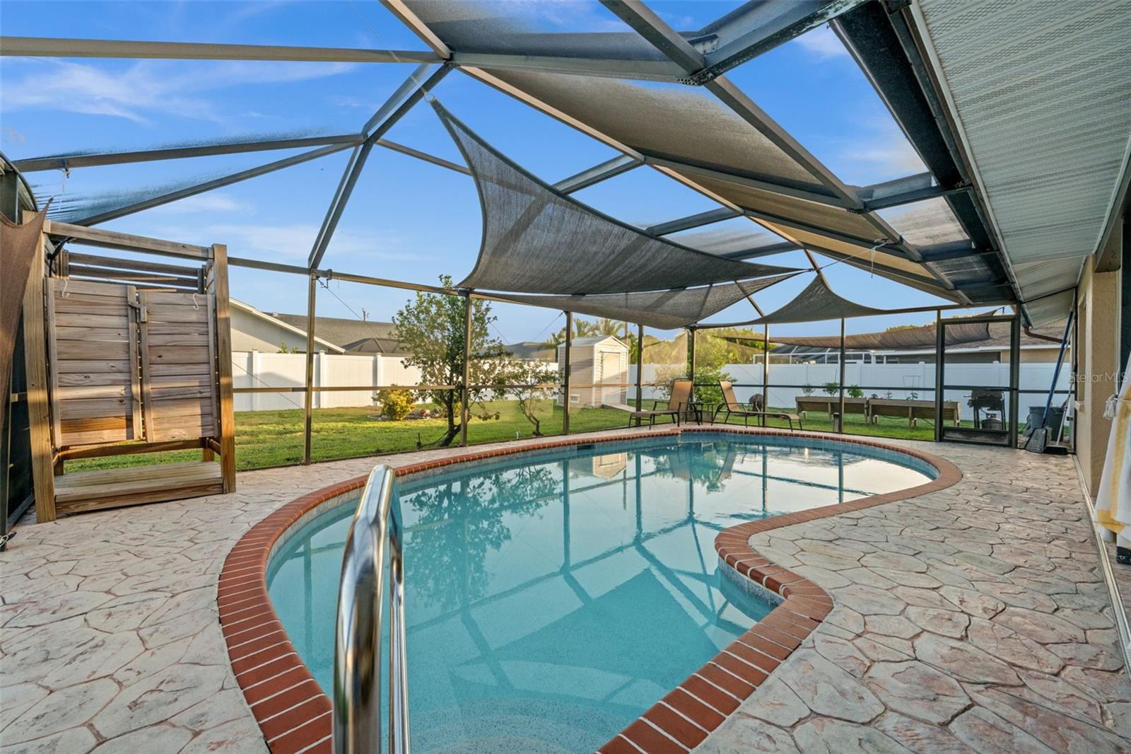 Screen-enclosed pool with brick-accented coping, stamped concrete deck, overhead sunshade canopy, and an outdoor shower enclosure.