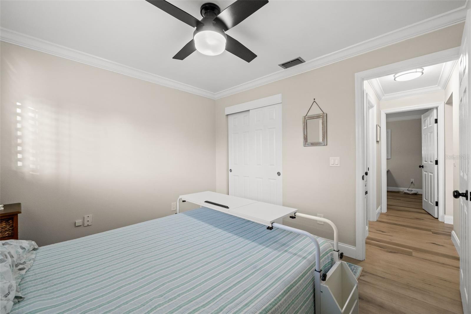 Neutral-toned interior featuring wood-finish flooring, crown molding, a central ceiling fan with integrated lighting, and white six-panel closet doors.