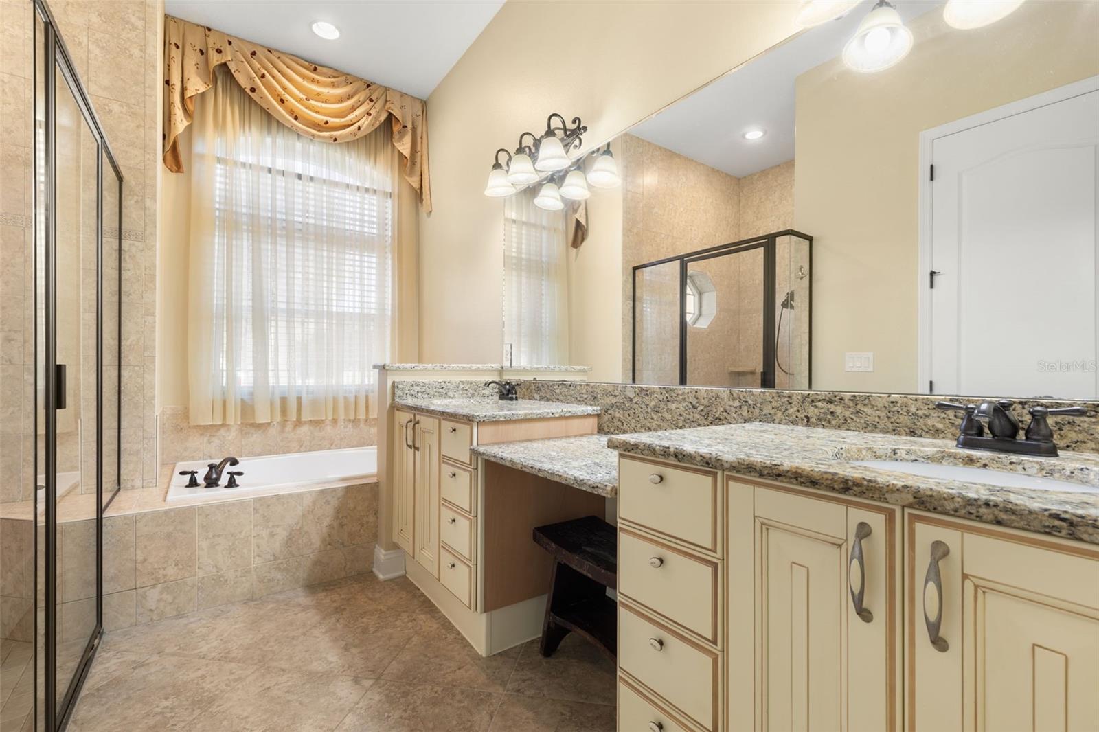 An Italian-inspired primary bathroom featuring dual vanities with granite countertops, a dedicated makeup area, soaking tub, and separate tiled shower.