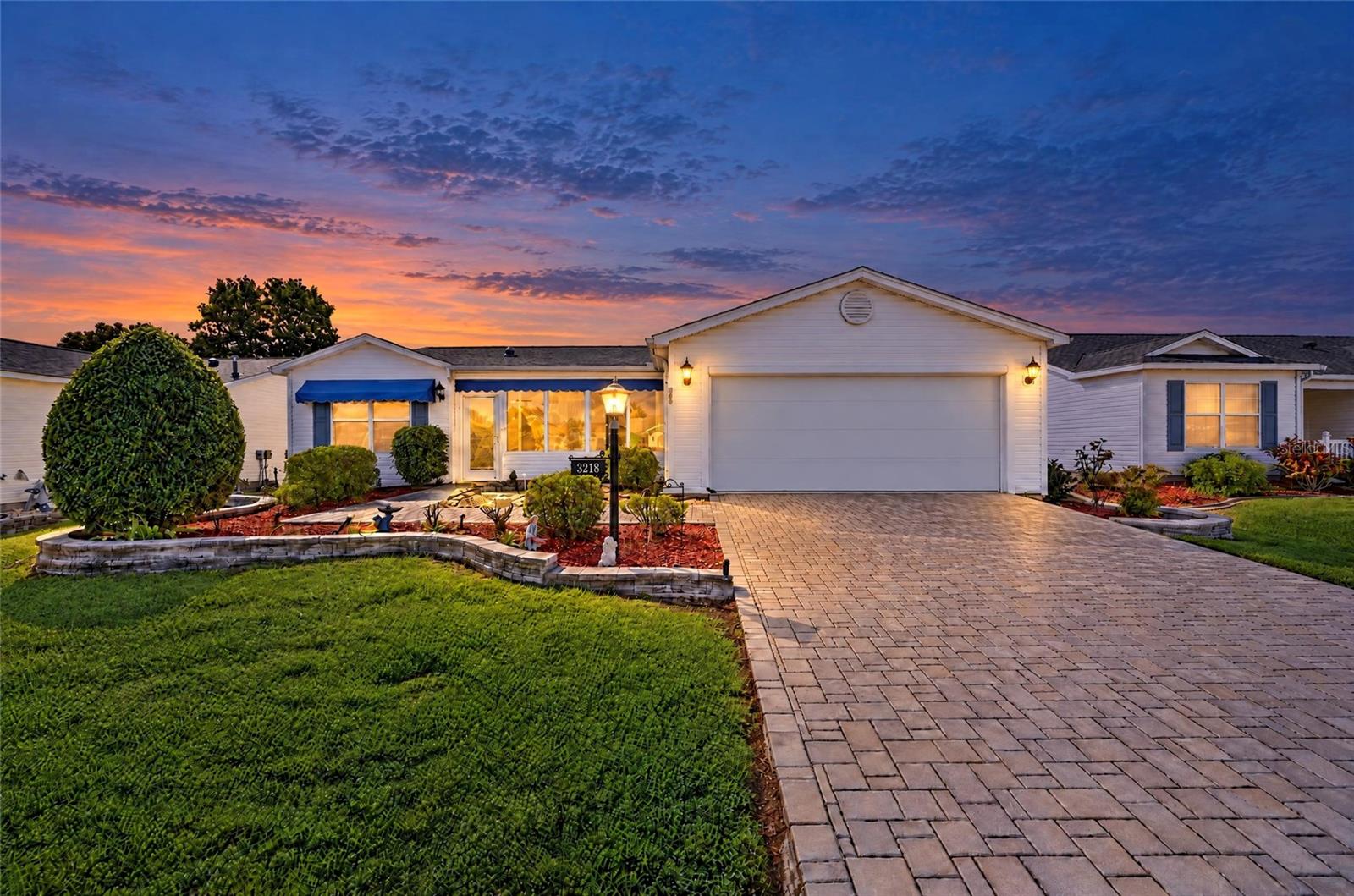 Beautiful twilight view showcasing the home's attractive landscaping, decorative stone retaining walls, expansive brick-paver driveway, blue awnings, and welcoming exterior lighting. Beautiful twilight view showcasing the home's attractive landscaping, decorative stone retaining walls, expansive brick-paver driveway, blue awnings, and welcoming exterior lighting.