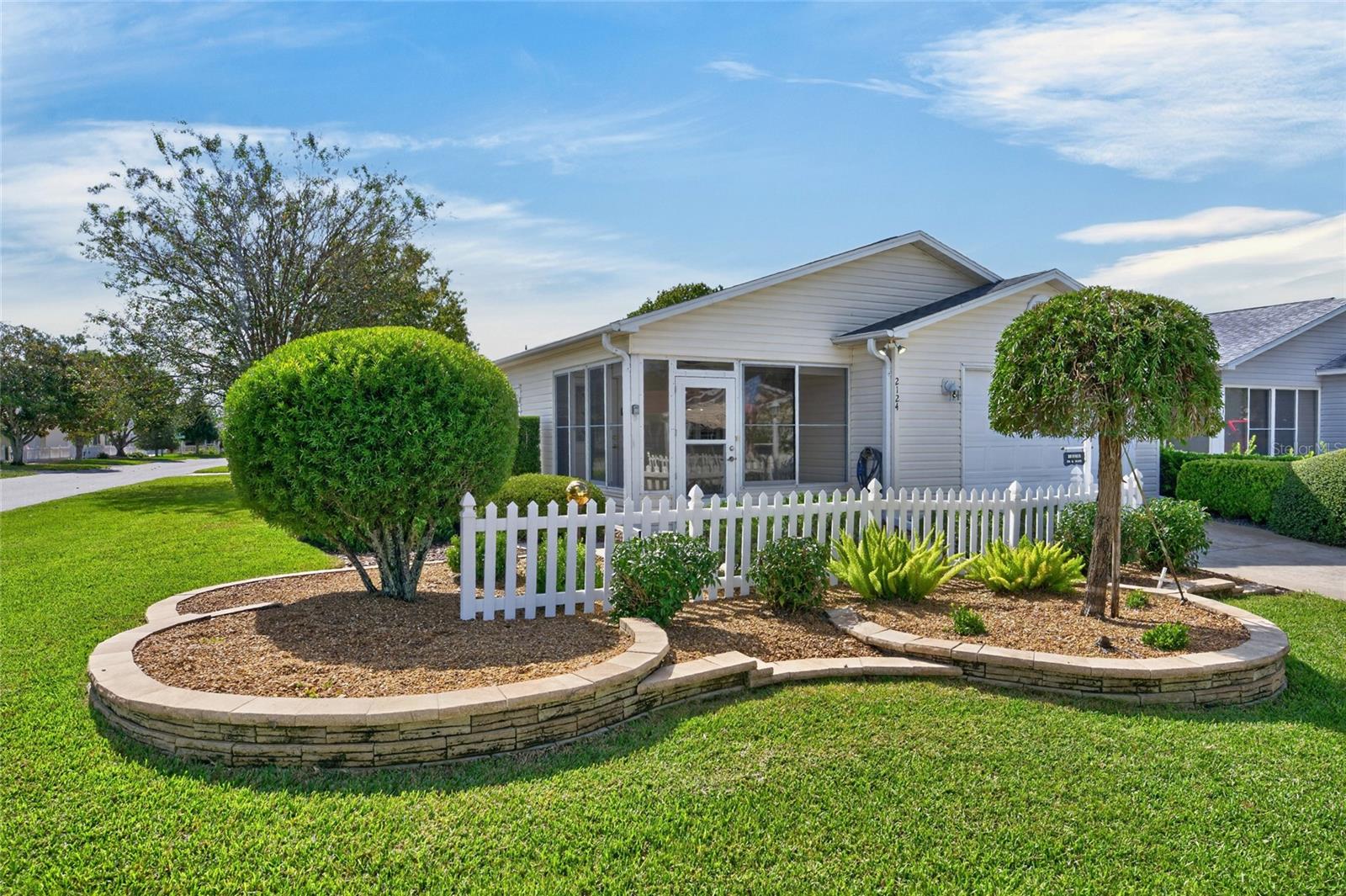 FRONT EXTERIOR - Elevated STACKED STONE landscape beds, manicured mature landscaping and a white PICKET FENCE frame the enclosed front lanai.