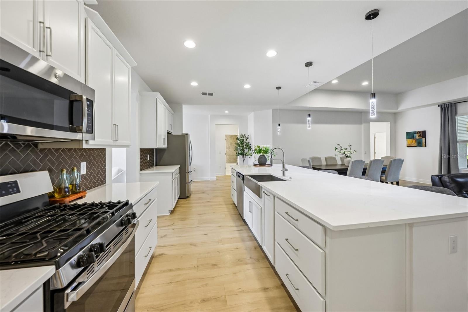 A massive QUARTZ ISLAND with bar seating sits beneath designer PENDANT LIGHTING, while STAINLESS STEEL APPLIANCES  including a GAS RANGE  pair perfectly with the stylish herringbone tile backsplash.