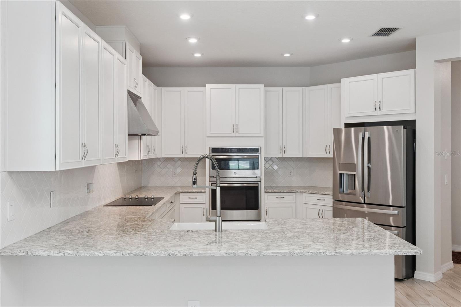 Kitchen  Spacious kitchen features abundant white cabinetry, granite countertops, a classic tile backsplash, stainless steel appliances, double wall ovens, and a generous peninsula with farmhouse-style sink and statement faucet.