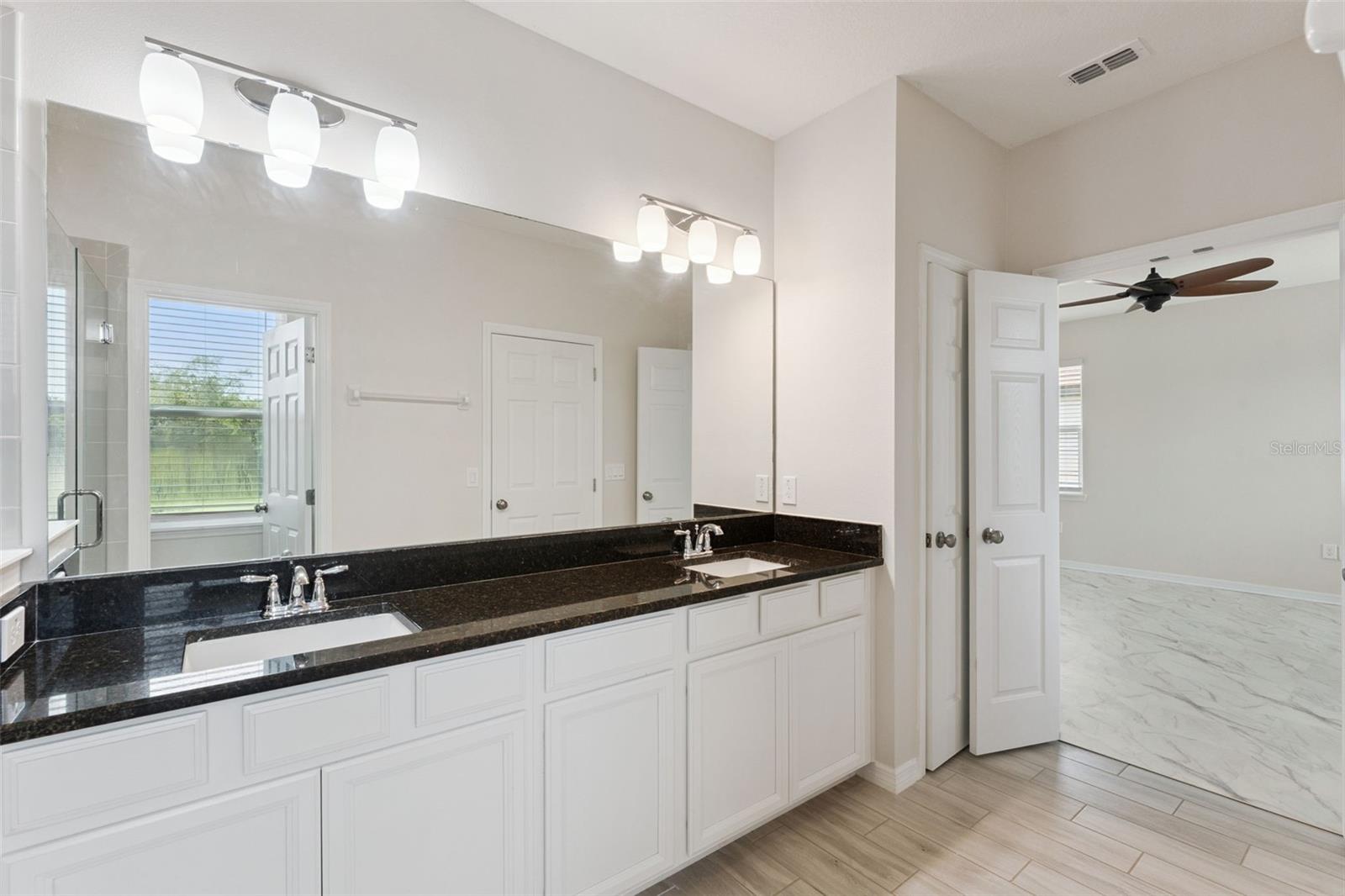 Primary Bathroom  Spacious primary bath features an extended dual-sink vanity with dark stone countertop, abundant cabinetry, a large wall mirror, and bright vanity lighting.