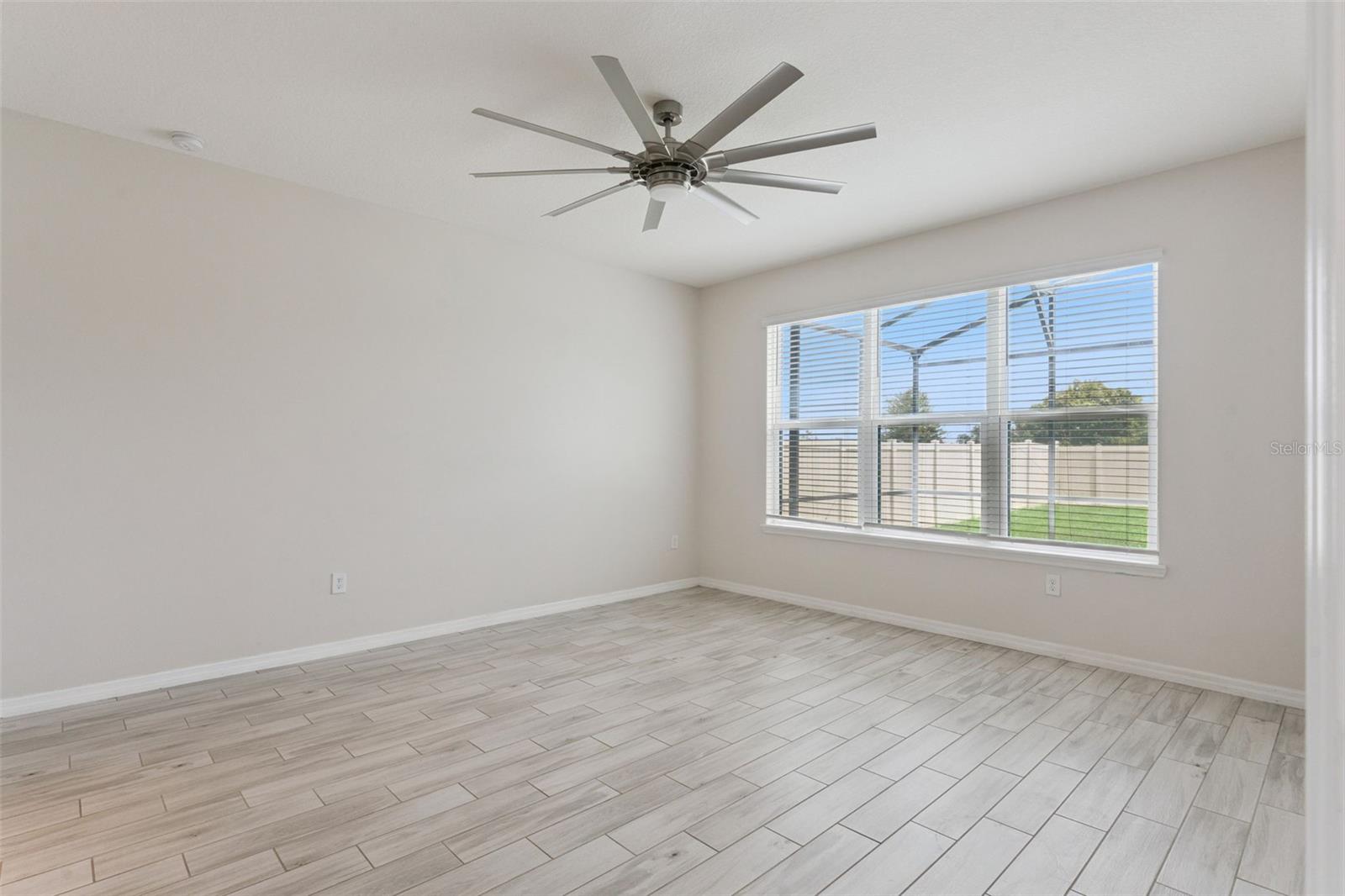 Primary Bedroom  Main-level primary bedroom offers generous proportions, wood-look tile flooring, a statement ceiling fan, and a wide window bringing in abundant natural light.