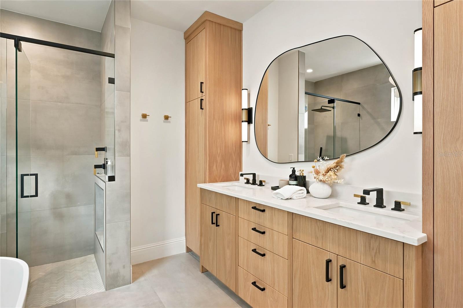 pairs warm wood cabinetry with clean contemporary finishes featuring a double vanity accented by mixed black and brass fixtures. A statement mirror & lighting add an artful touch to the serene, sophisticated space.