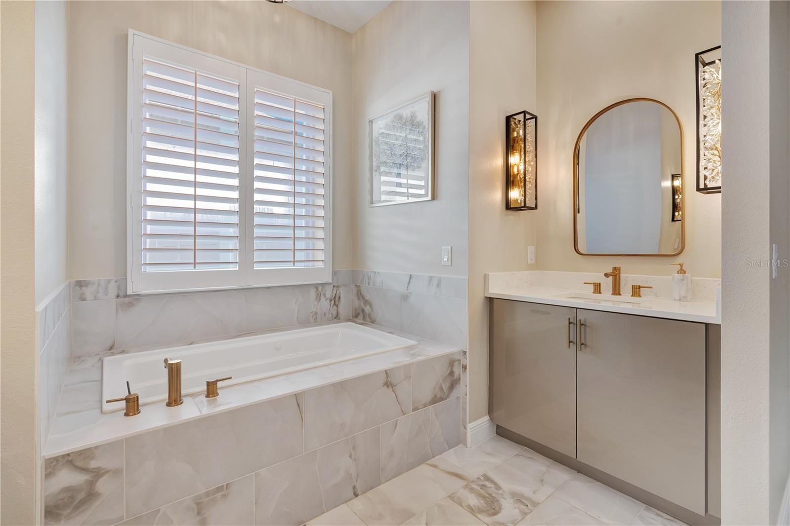 Relaxing bath retreat with a soaking tub beneath plantation shutters, decorative mirror and gold-tone fixtures.