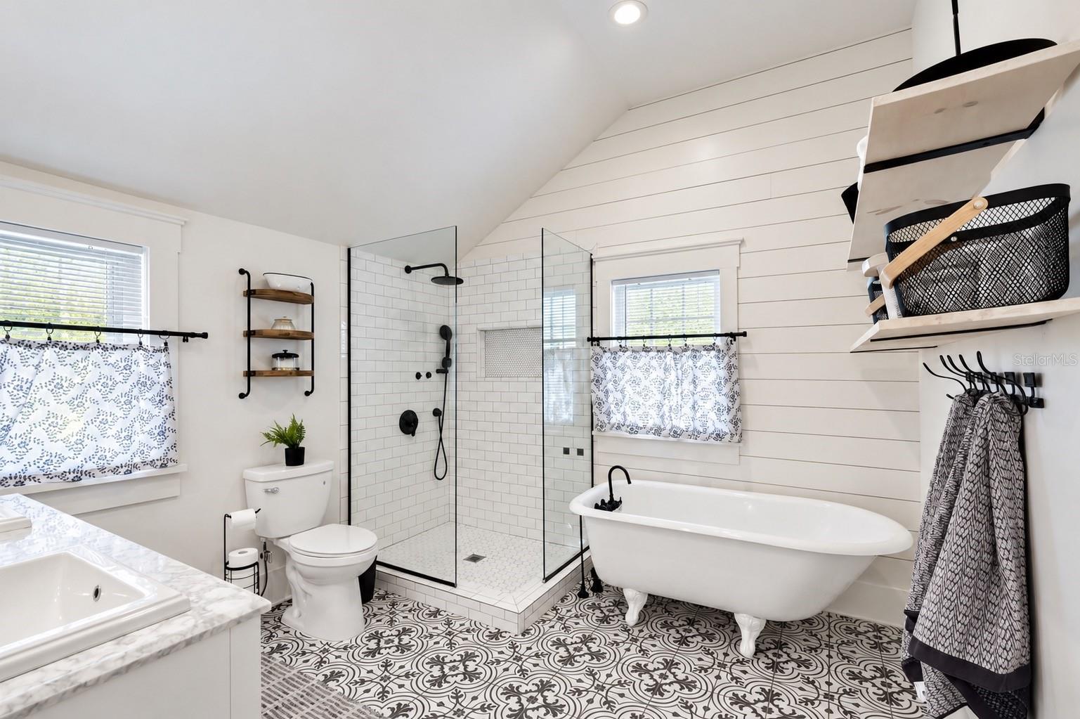 Beautifully updated upstairs bathroom conveniently located between both bedrooms, featuring a charming clawfoot soaking tub, spacious glass-enclosed walk-in shower, stylish patterned tile flooring, shiplap walls, and elegant black fixtures.