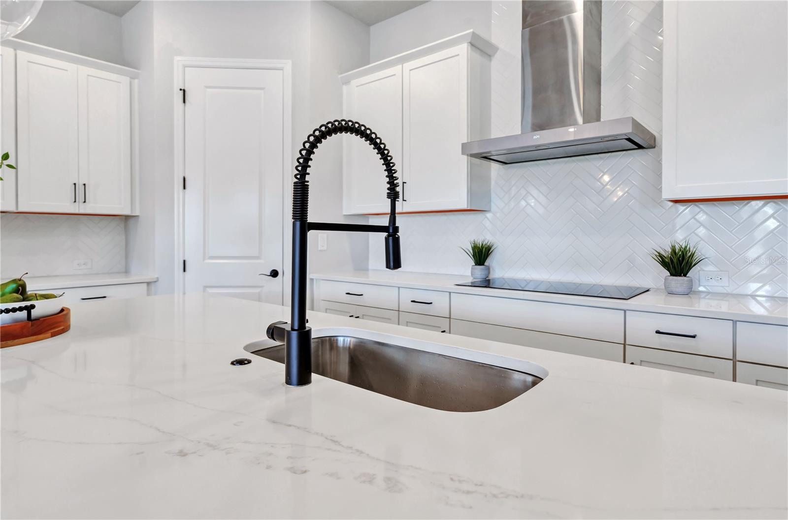 Quartz counters, undermount sink, matte black faucet, and herringbone tile backsplash with a view into the home.