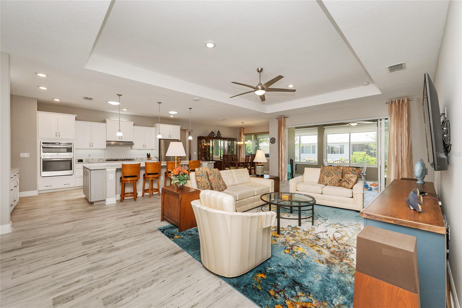 Spacious open-concept great room featuring a tray ceiling, recessed lighting, ceramic plank flooring, and stacking sliding glass doors with a zero-threshold entry to the enclosed lanai, creating a seamless transition between indoor and outdoor living spaces.