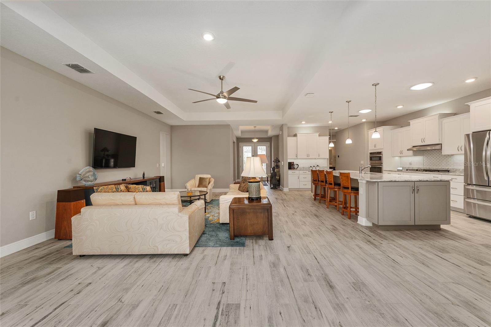 A stunning view of the spacious great room and beautifully appointed kitchen, showcasing an elegant tray ceiling, ceramic plank flooring, recessed lighting, and two kitchen solar tubes that bring additional natural light into the space.