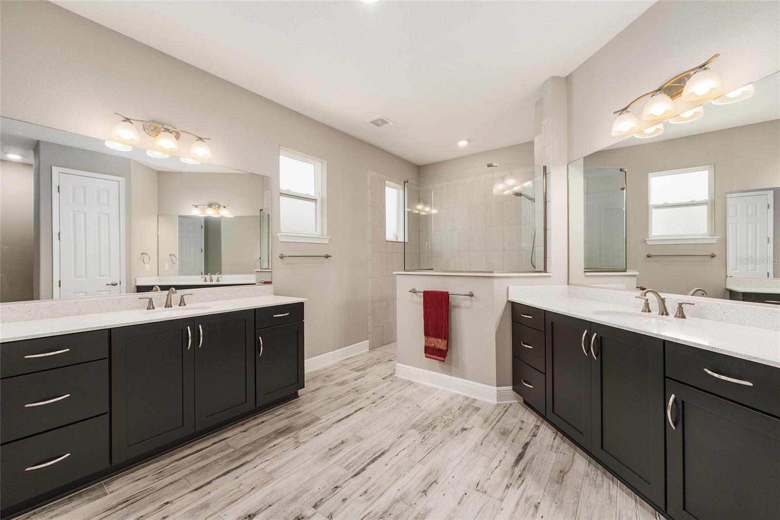 Spacious first primary ensuite bathroom featuring separate vanities with generous countertop space, abundant cabinet and drawer storage, ceramic plank flooring, and a large walk-in shower with glass enclosure.