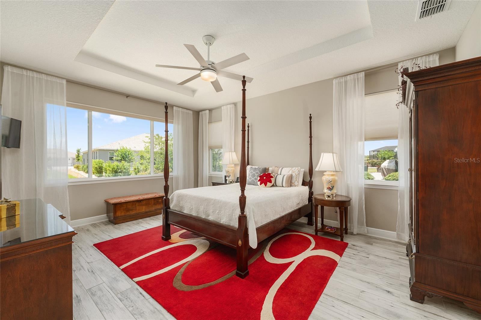 Spacious first primary suite, currently used as the owner's suite, featuring an elegant tray ceiling, ceiling fan with integrated lighting, ceramic plank flooring, neutral finishes, and multiple windows that fill the room with natural light. Wall-mounted TV included in the sale.