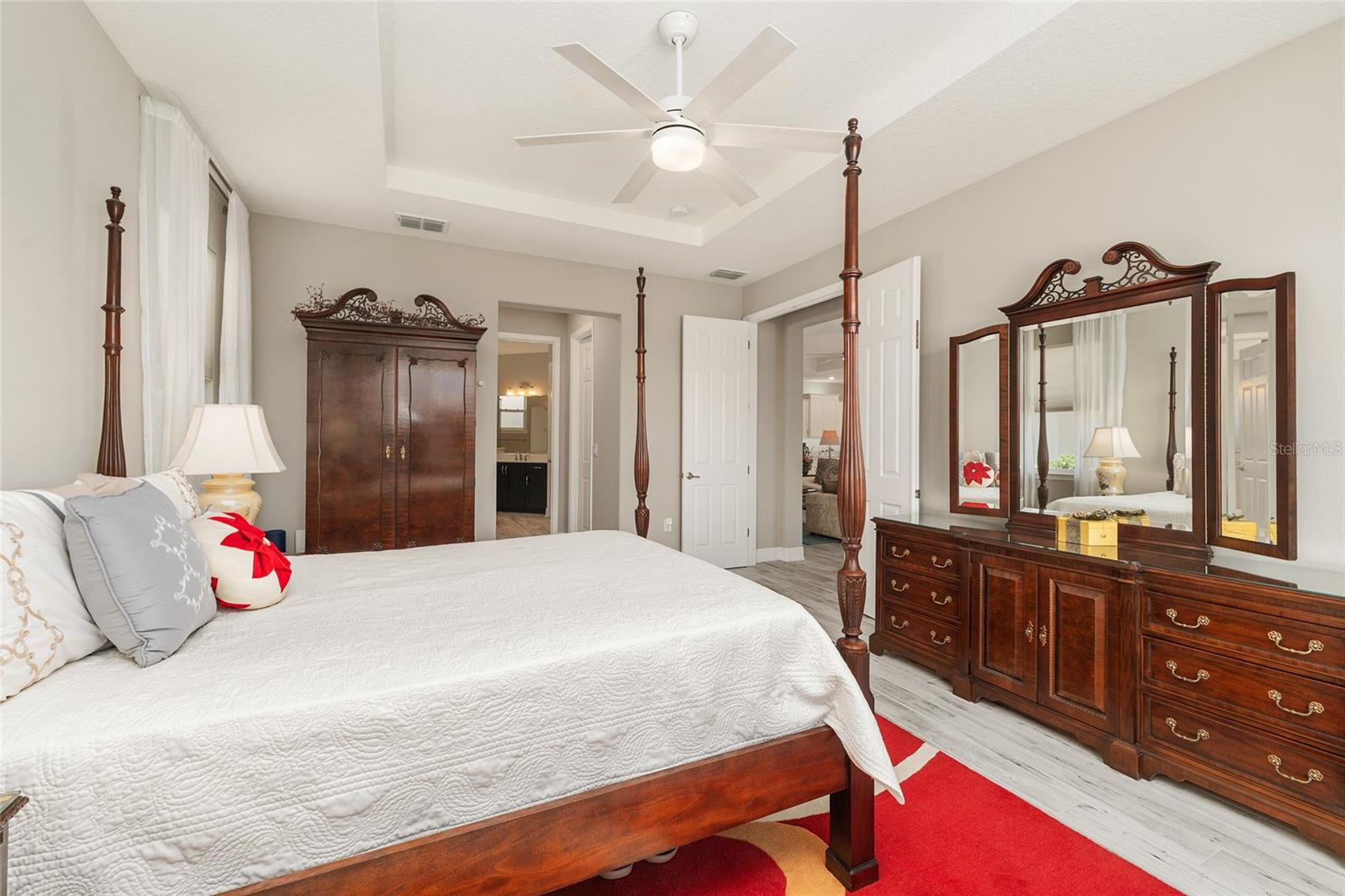 Another view of the first primary suite, currently used as the owner's suite, featuring a double-door entry, tray ceiling, ceramic plank flooring, and convenient access to the ensuite bathroom.