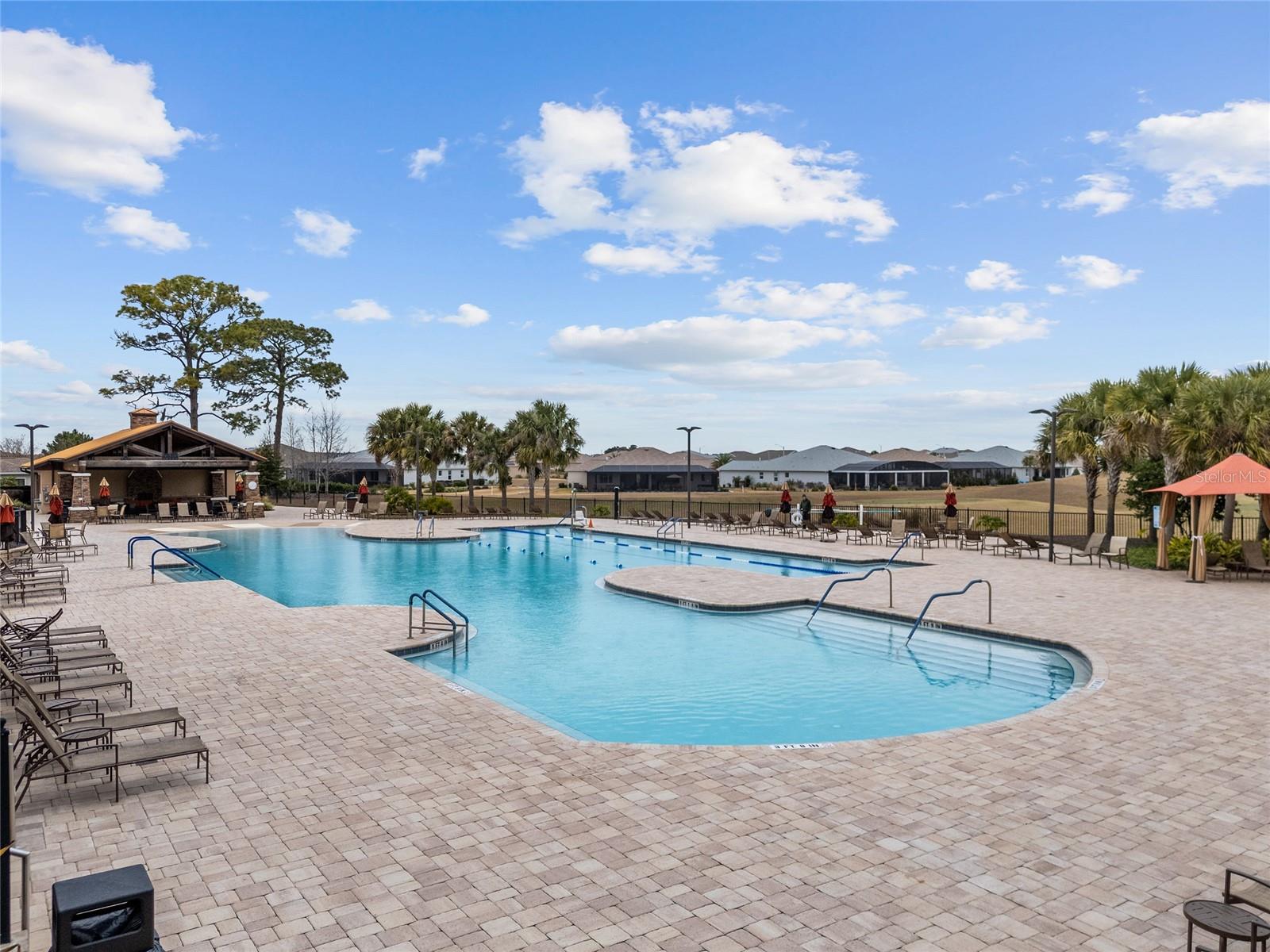 Another beautiful view of the Candler Hills resort-style heated pool, showcasing the expansive sun deck, abundant lounge seating, tropical landscaping, and covered pavilionan inviting setting for soaking up the Florida sunshine and socializing with neighbors.