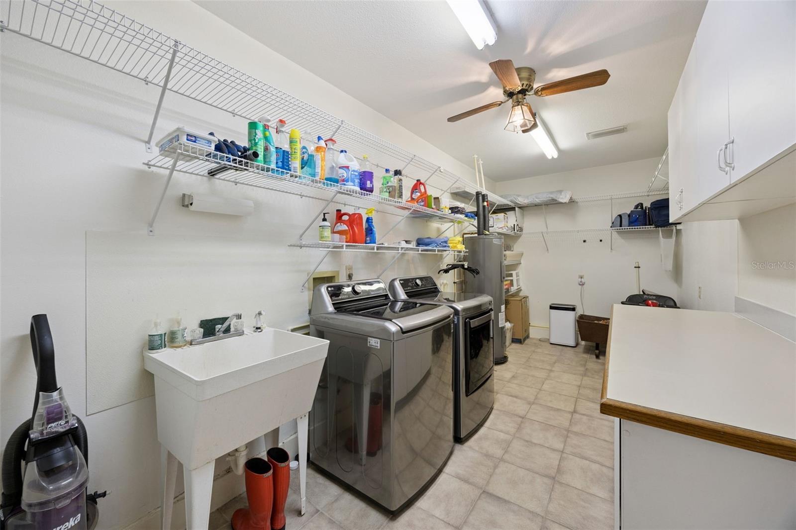Dedicated Laundry Room: Large utility room featuring a laundry sink, side-by-side washer and dryer, water heater, overhead storage wire racks, and upper cabinets.
