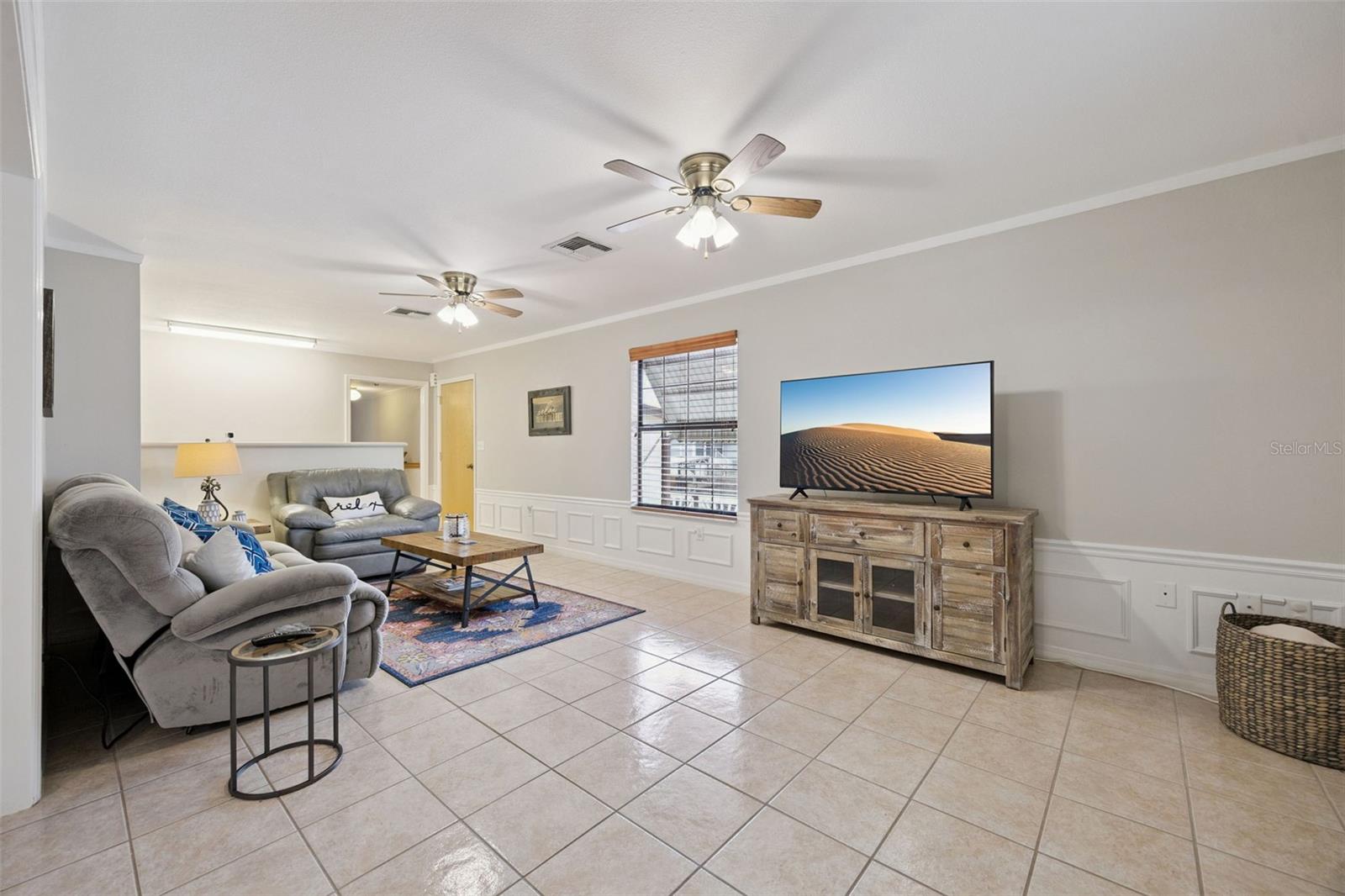 Main Living Room: Expansive open living room featuring neutral ceramic tile flooring, classic wainscoting trim, ceiling fans, and built-in media area.