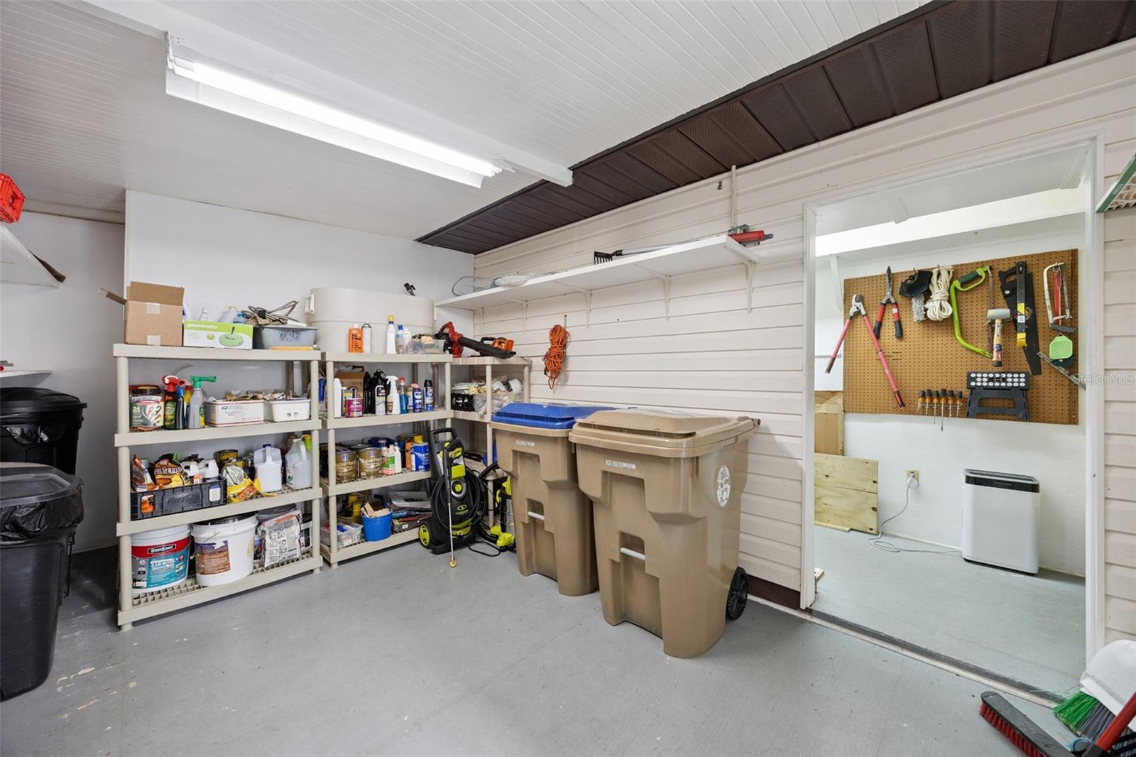 Workshop & Storage Utility Room: Organized storage space featuring built-in shelving, concrete flooring, overhead fluorescent lighting, and an adjacent workshop area with pegboard tool organization.