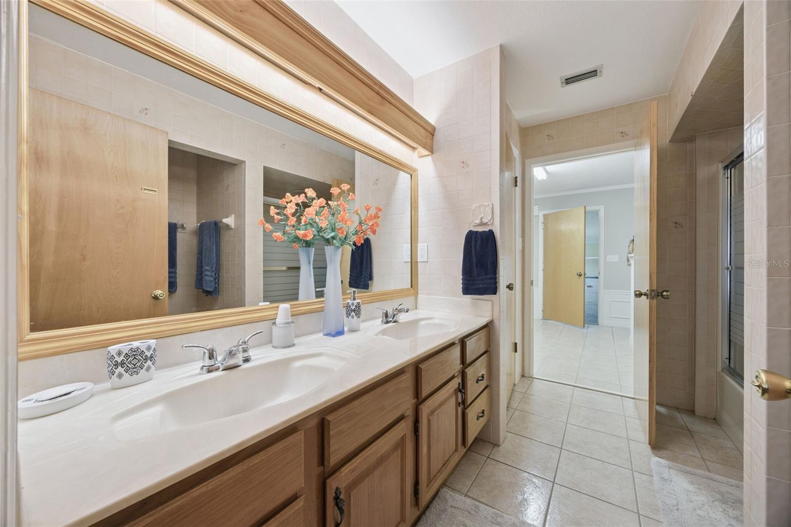 Dual Sink Primary Vanity: Long primary bathroom vanity with double sinks, warm wood cabinetry, full-width mirror, and integrated lighting.