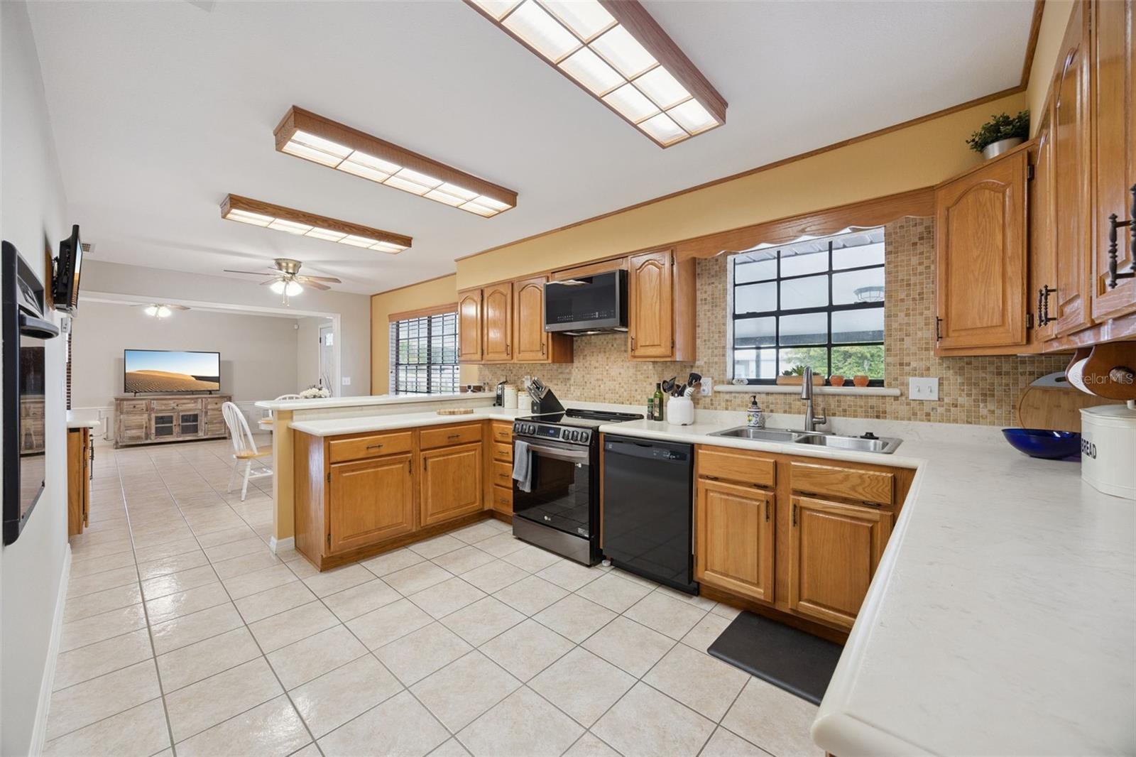 Functional Kitchen Layout: Bright kitchen equipped with traditional oak cabinets, mosaic backsplash, black appliances, double sink under a window, and continuous tile flooring.