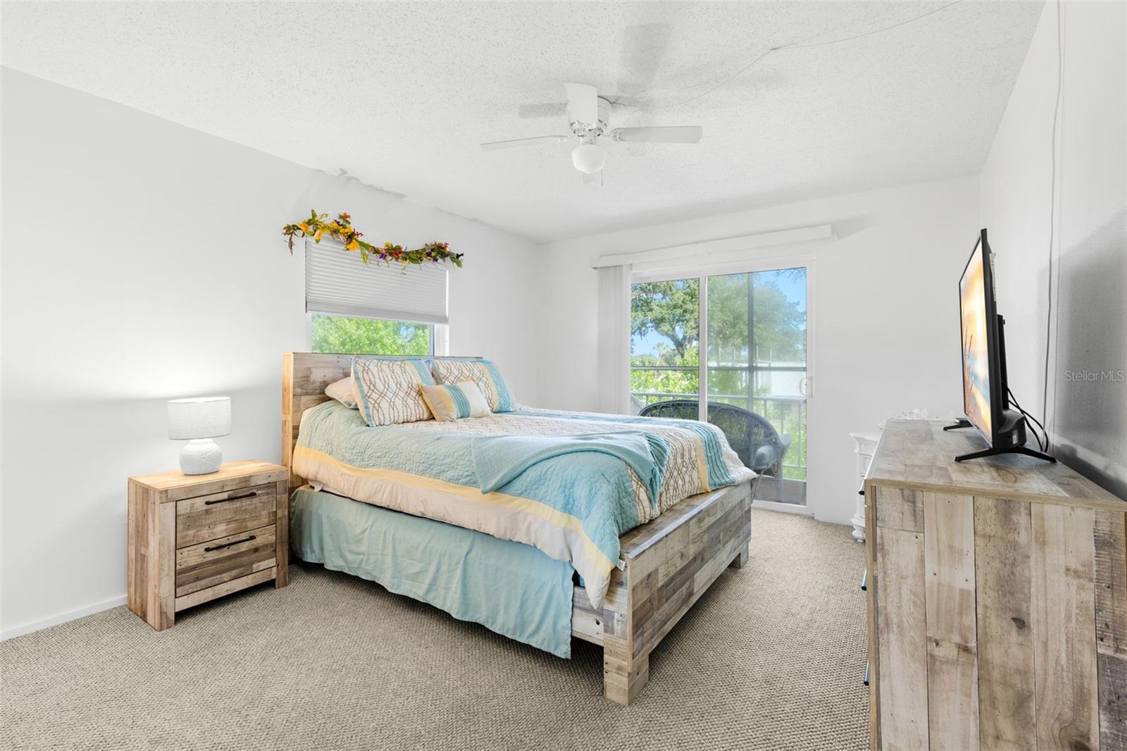A neutral backdrop of light walls and carpet keeps the primary bedroom bright, while a second window and sliding glass doors bring in natural light and frame the greenery outside.