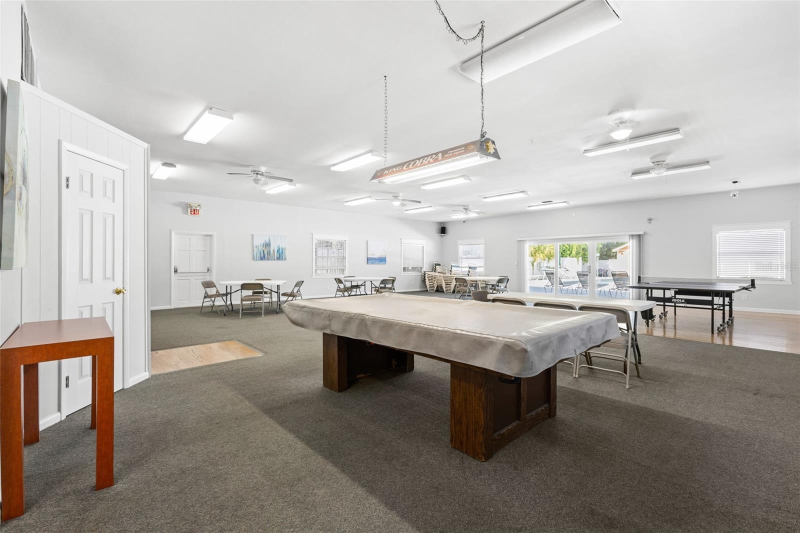 The clubhouse offers a large gathering space with billiards, ping-pong and multiple tables for games, cards or community events, with doors opening toward the pool area beyond.