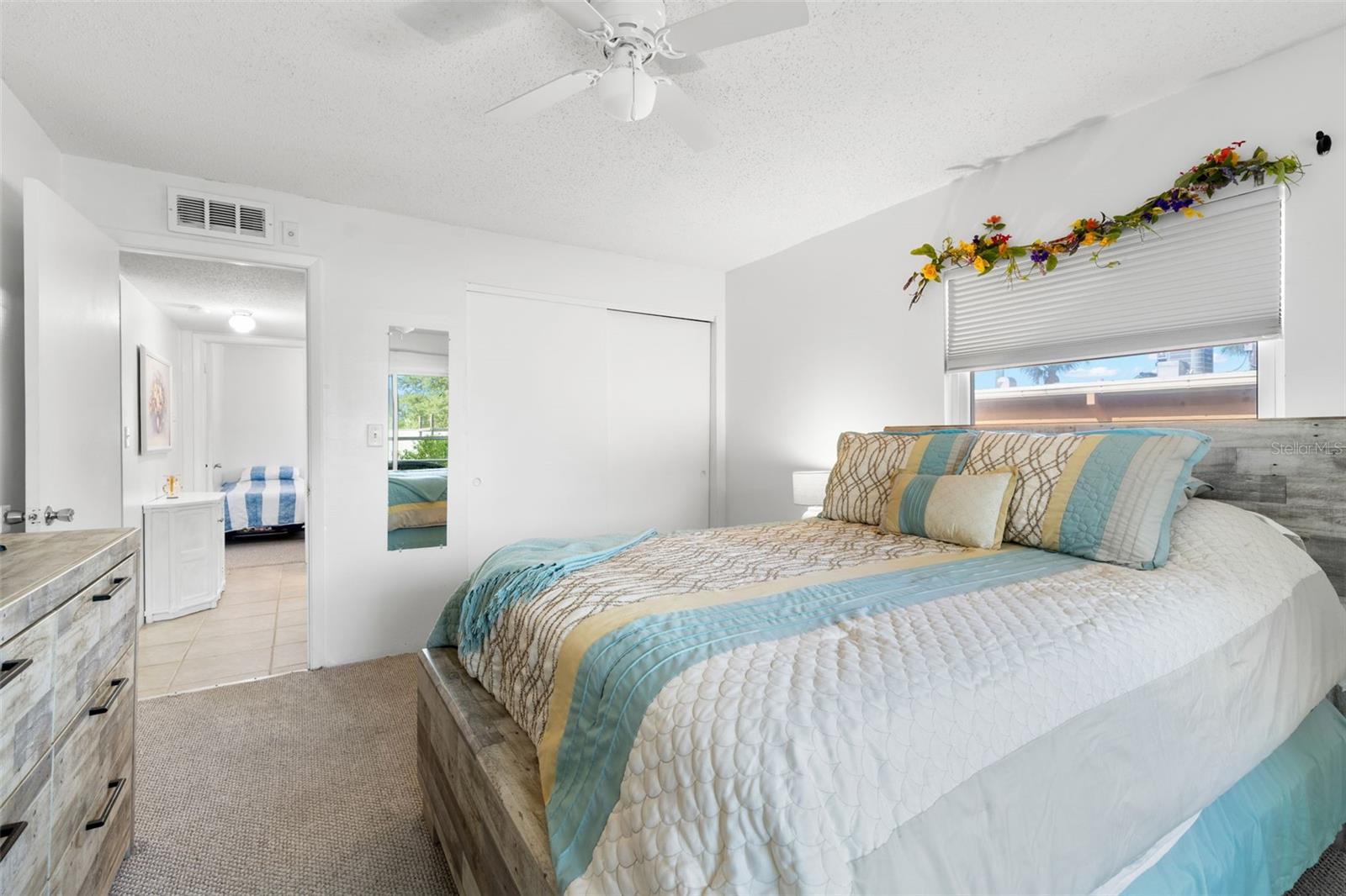 A built-in closet with sliding doors provides storage in the primary bedroom, while neutral carpet, a ceiling fan and a window complete the space.