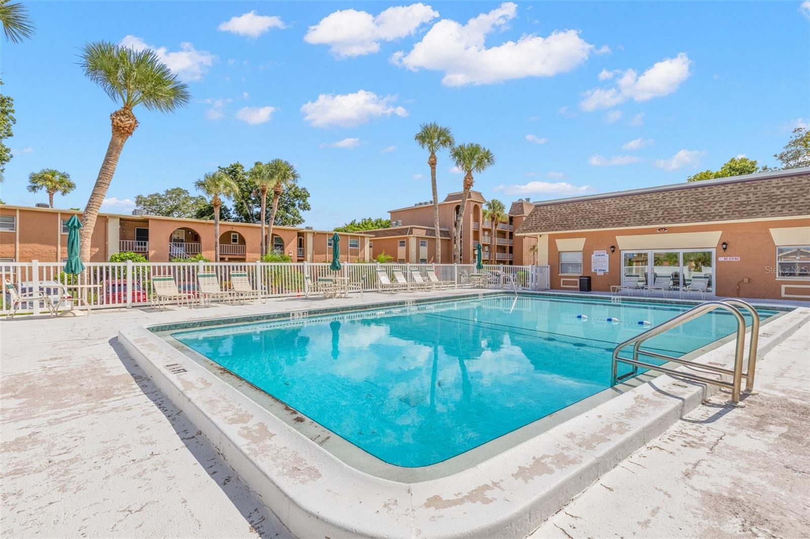 The community pool is surrounded by a spacious deck with lounge seating and umbrellas, while palms and the neighboring clubhouse create an inviting spot to relax outdoors.