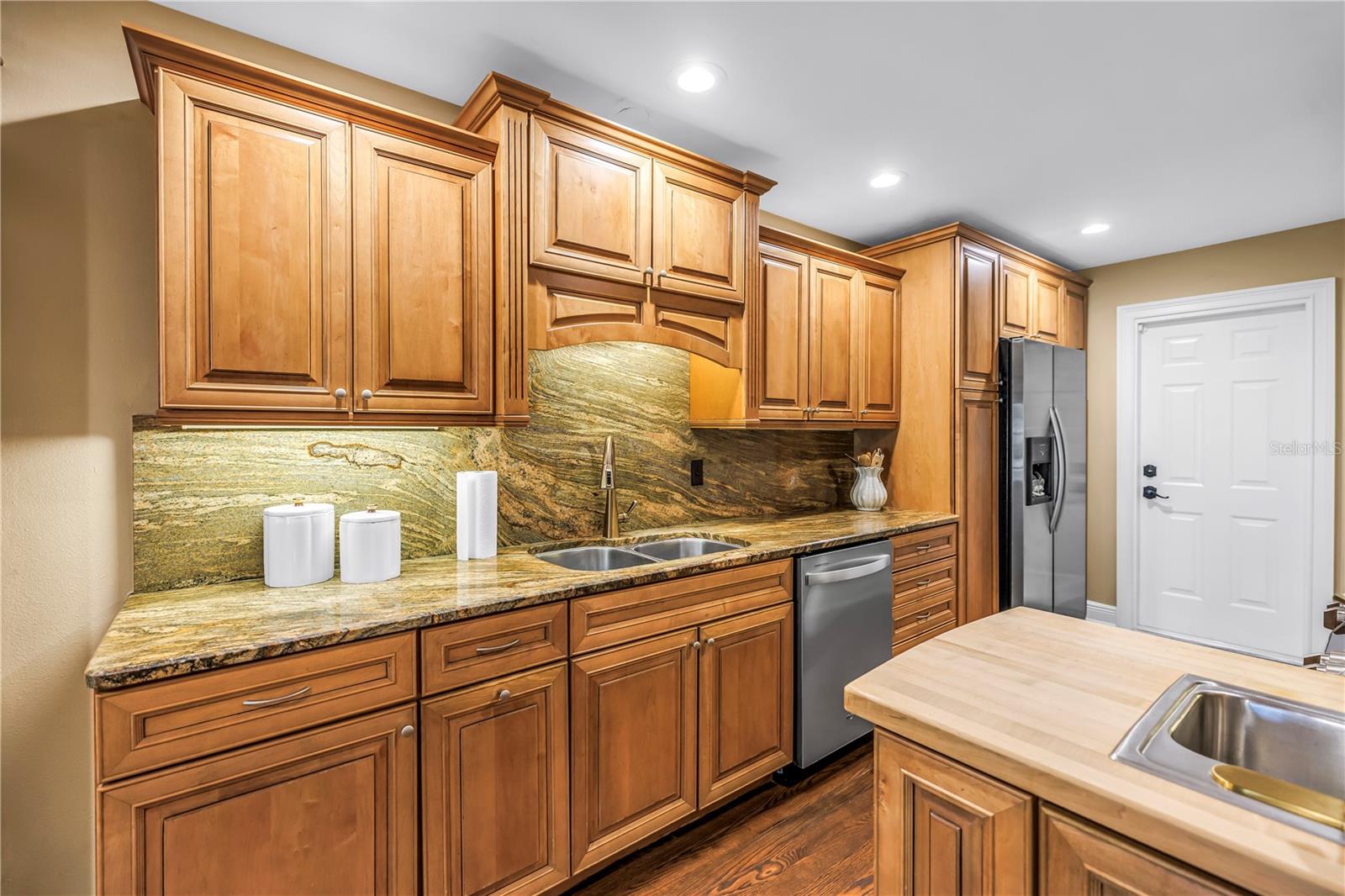 This custom kitchen features solid wood cabinetry, granite countertops, and a matching backsplash. It is equipped with updated stainless steel appliances, a chopping block island, and an integrated prep sink. An ideal space for any culinary enthusiast.