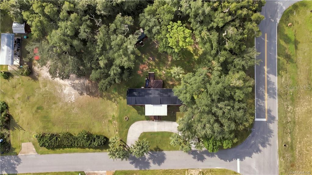 Aerial Property View  Beautiful overhead perspective showcasing the .29-ACRE CORNER HOMESITE, METAL ROOF, circular concrete driveway, mature shade trees & spacious lawn.