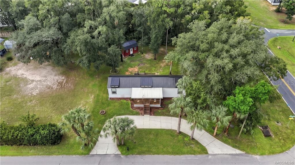 Aerial Property View  A beautiful overhead perspective of the .29-ACRE CORNER HOMESITE, showcasing the METAL ROOF, COVERED FRONT PORCH, circular concrete driveway, mature trees & detached storage building.