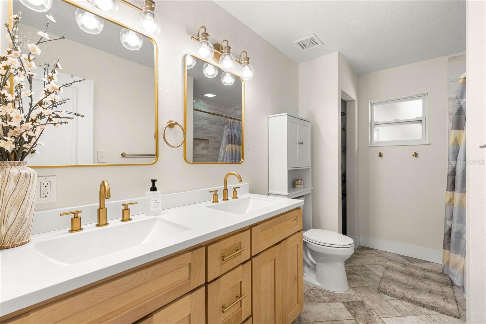 The stylish second bathroom features a double-sink vanity with natural wood cabinetry, crisp white countertops, brushed gold fixtures, coordinating mirrors, and contemporary glass-globe lighting. Neutral tile flooring and a tub/shower combination complete this thoughtfully updated space.