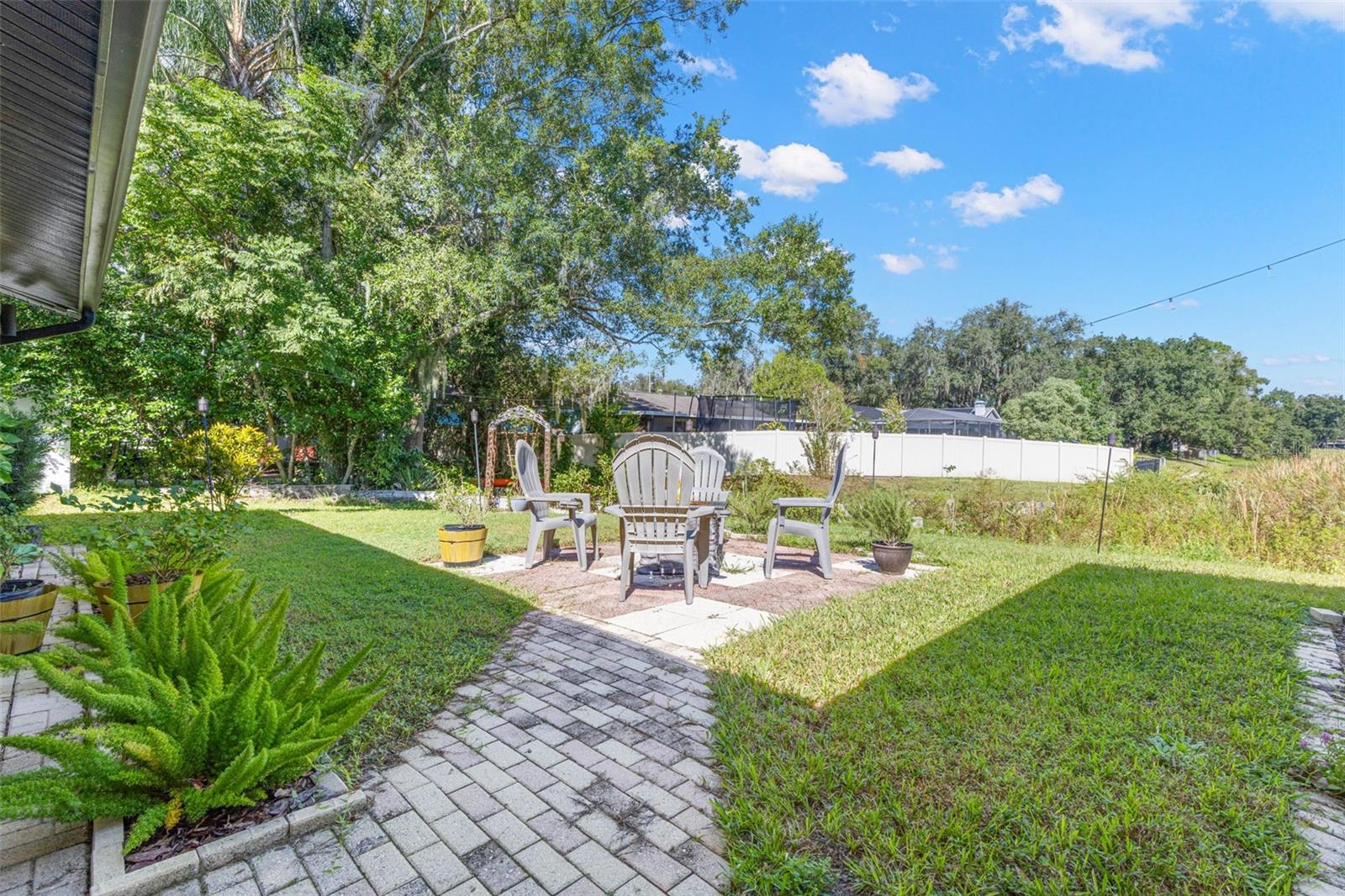 A charming paver walkway leads to an inviting outdoor seating area, perfect for morning coffee, evening conversations, or gathering around a fire pit. Mature trees, lush landscaping, and an open lawn create a peaceful backyard retreat just steps from the pond.