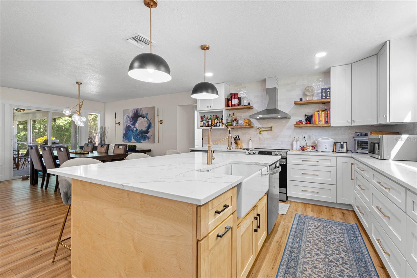 A chef-inspired kitchen with an apron-front farmhouse sink, professional-style gold faucet, convenient pot filler above the range, stainless steel range hood, and floating wood shelves. The oversized island provides additional cabinetry and generous workspace, while the open layout connects effortlessly to the dining area.