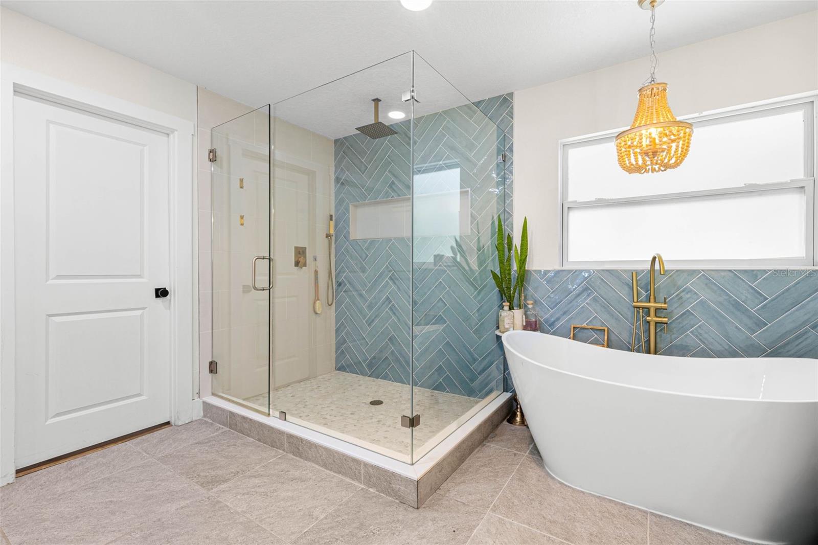 A freestanding soaking tub takes center stage beneath an elegant gold chandelier, complemented by a floor-mounted gold tub filler and beautiful blue-green herringbone tile. The adjoining glass-enclosed shower completes this luxurious, spa-inspired retreat.